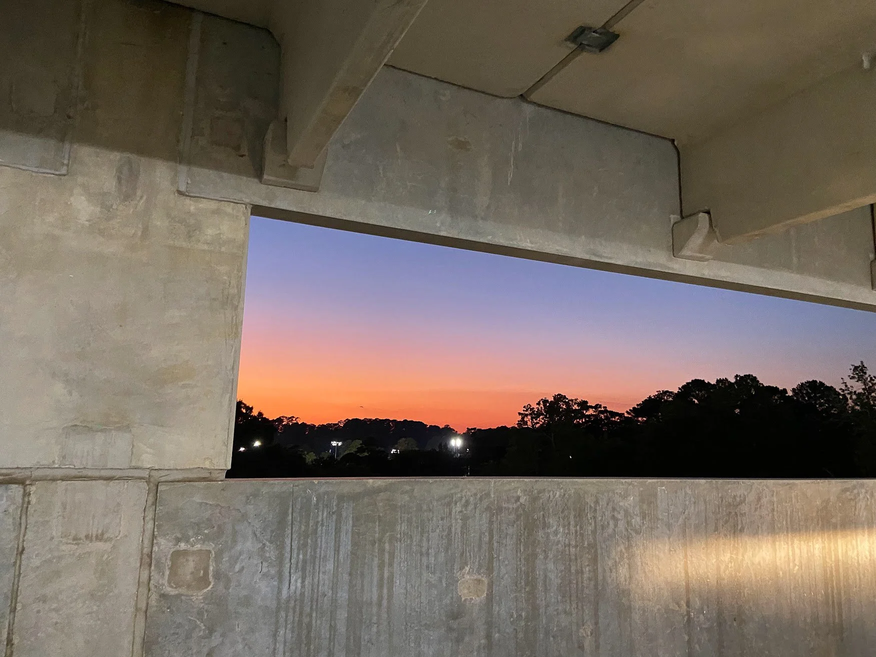 Sunset view through an unfinished concrete wall with trees and lights in the distance.