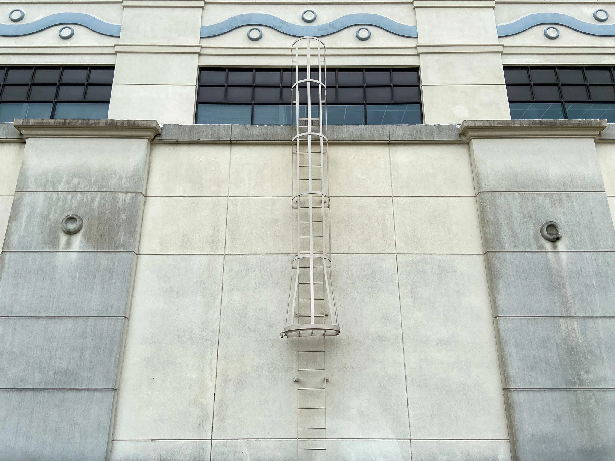 Exterior of a building with a metal fire escape ladder attached to a beige wall, above a row of dark window panes.