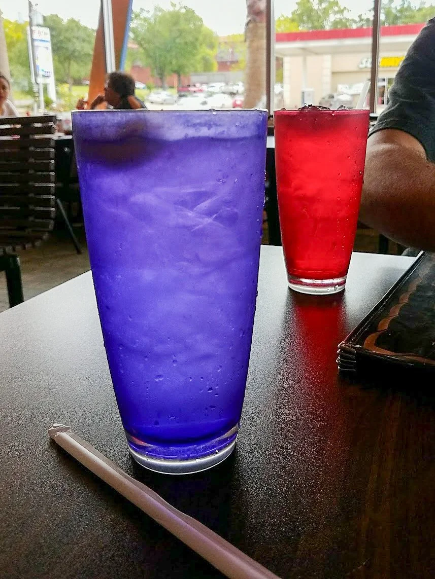Two glasses filled with cold beverages, one purple and one red, on a black table inside a restaurant with large windows overlooking a parking lot and greenery.