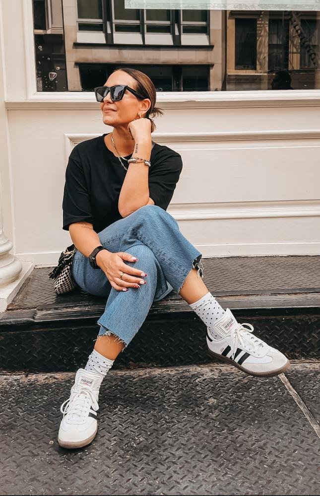 Woman sitting on steps wearing sunglasses, a black shirt, blue jeans, white sneakers with stripes, and polka dot socks.