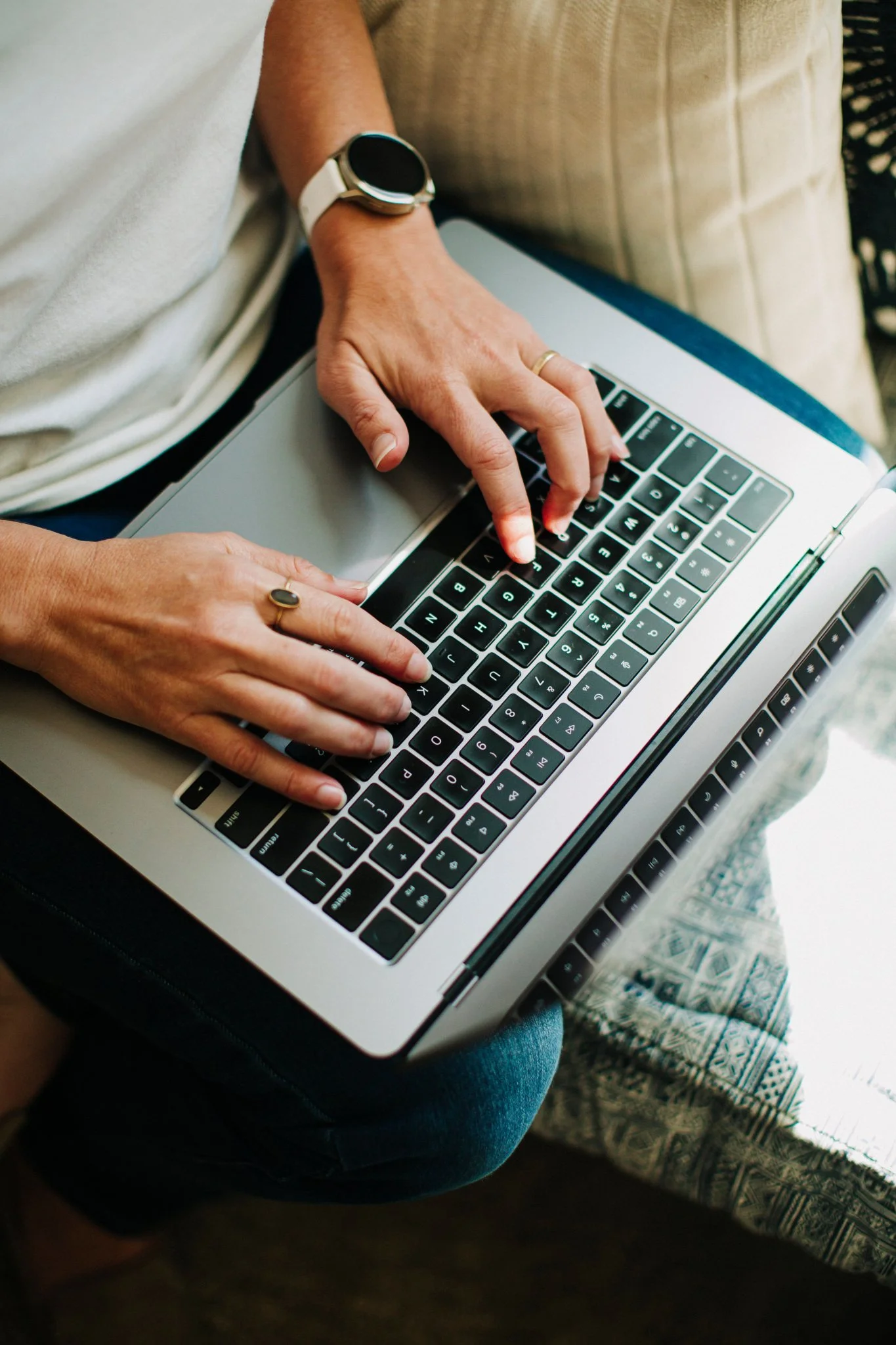 A person wearing a white watch and a ring on their left hand using a silver laptop with black keys.