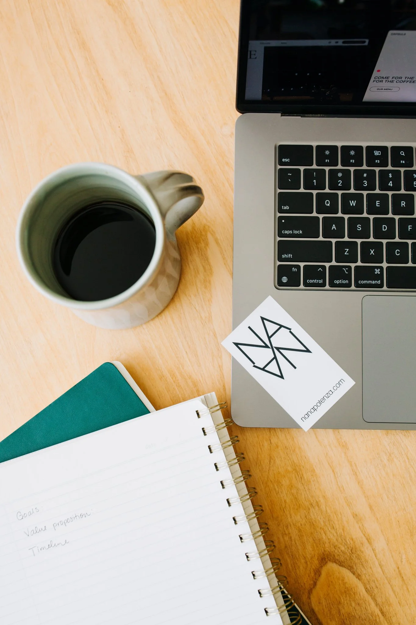 A workspace on a wooden desk with a laptop, a mug filled with black coffee, an open notebook with handwritten notes, and a business card with Nana's logo and website URL.