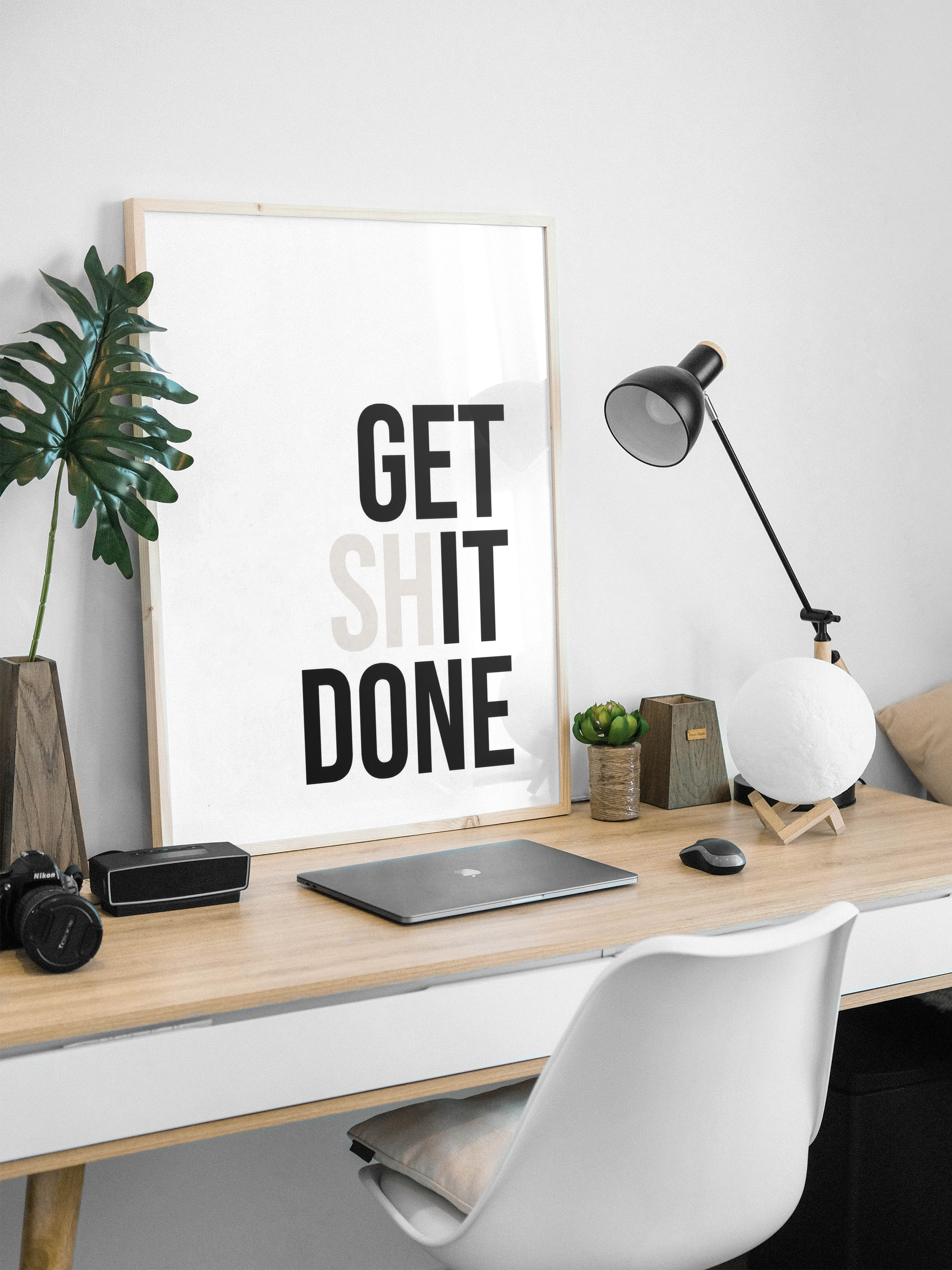 A minimalist workspace with a wooden desk and a framed poster that reads 'Get Shit Done' in black and beige text.
