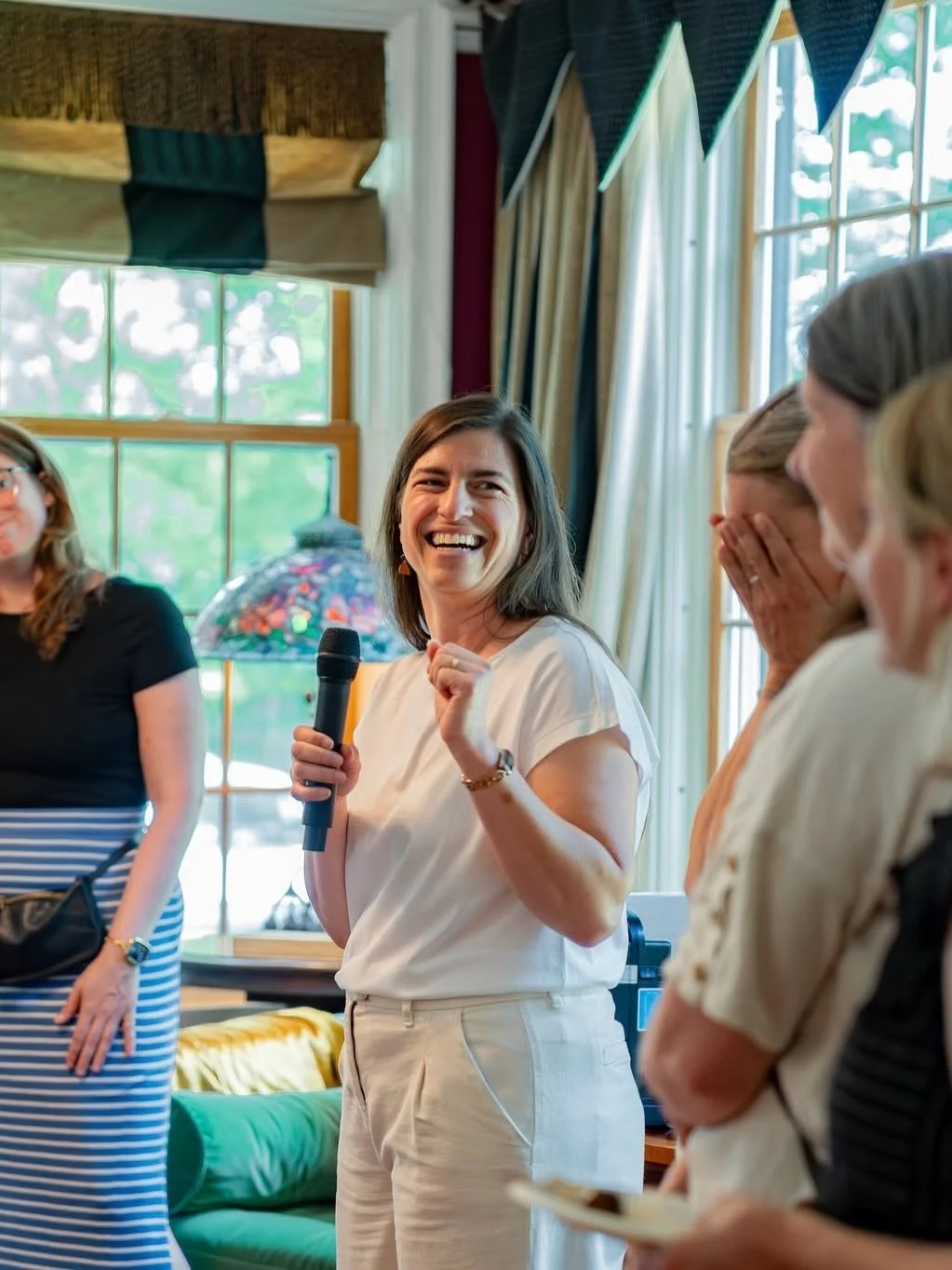 Nana Potenza holding a microphone, smiling, and speaking in a room with large windows and colorful curtains surrounded by a group of women, some covering their mouths and smiling.