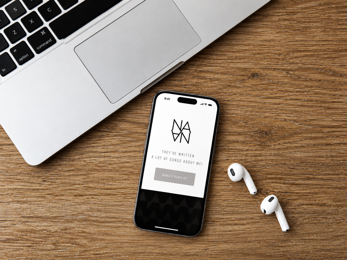 A laptop, a smartphone displaying a music app, and a pair of wireless earbuds on a wooden desk.