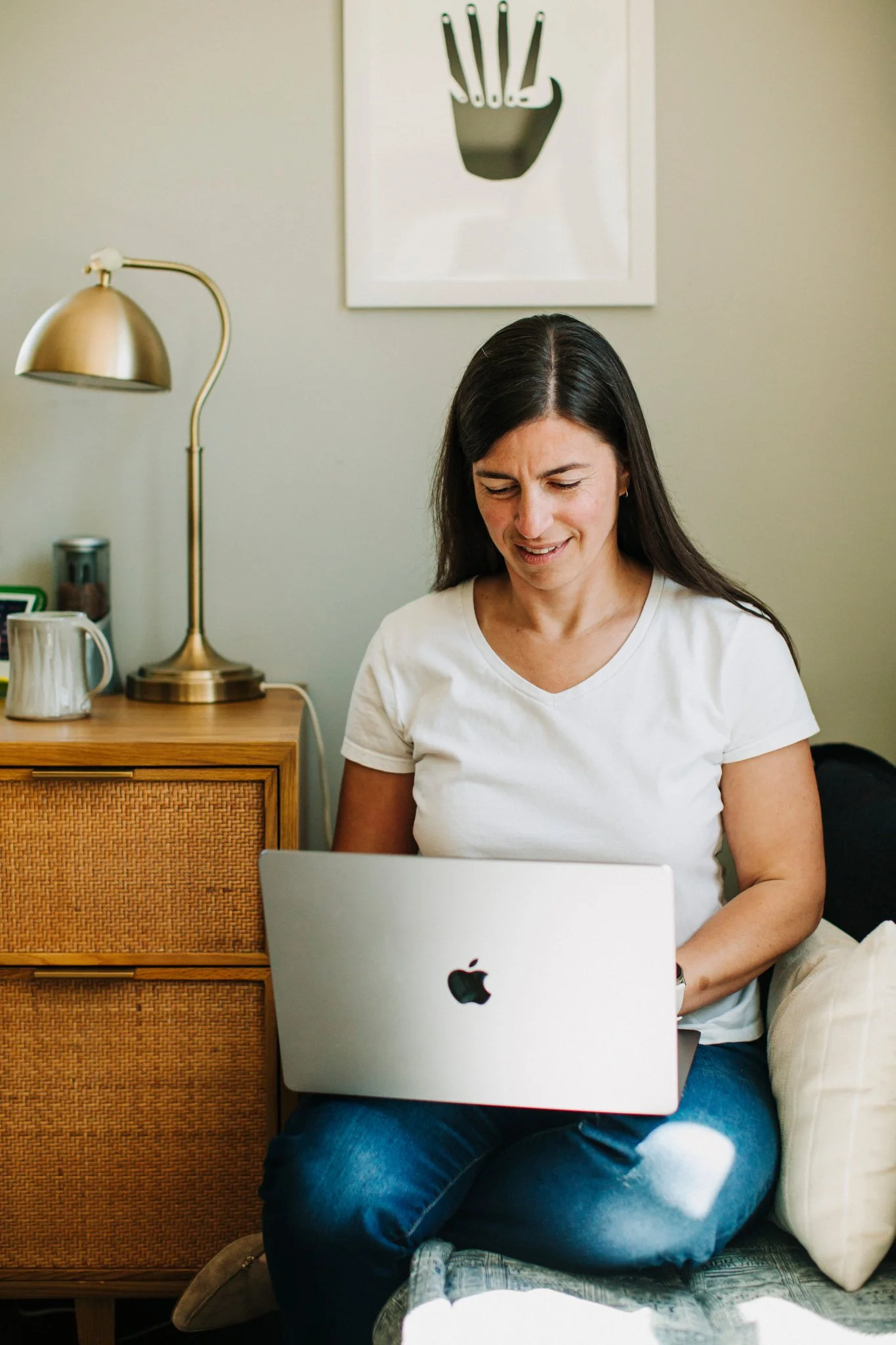 Nana Potenza with long dark hair sitting on a day bed, using a silver MacBook.