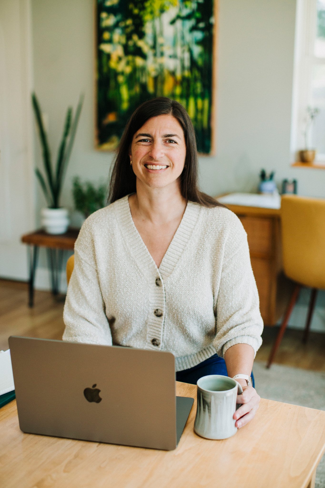 Nana Potenza with dark hair and a beige sweater sitting at a desk in a cozy, well-lit room with a laptop and a coffee mug, smiling at the camera.