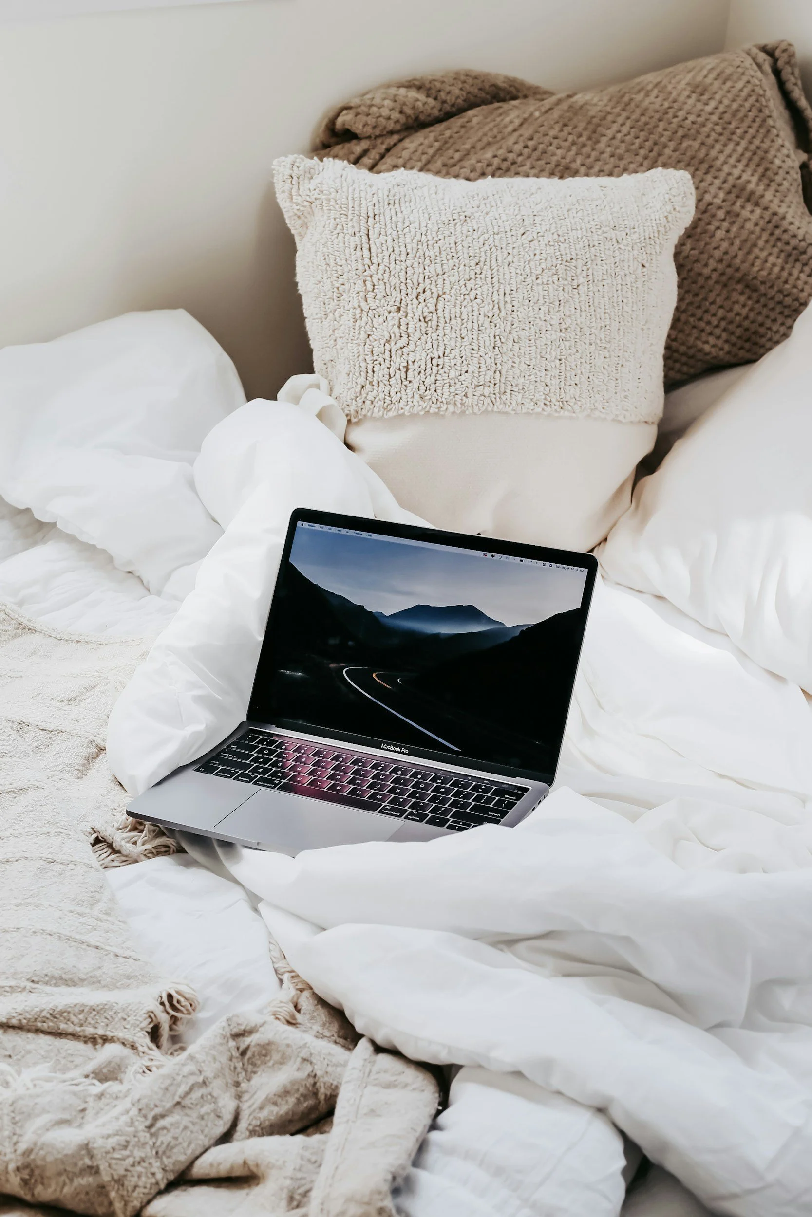 Stock photo of a laptop on a bed, surrounded by beige and brown throw blankets and pillows.