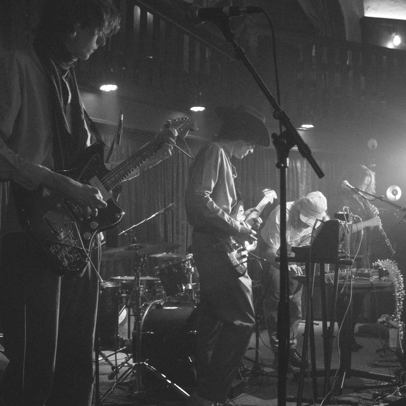 A black and white photo of a band performing on stage, with multiple guitar players, a drummer, and a keyboardist, with stage lighting and microphones.