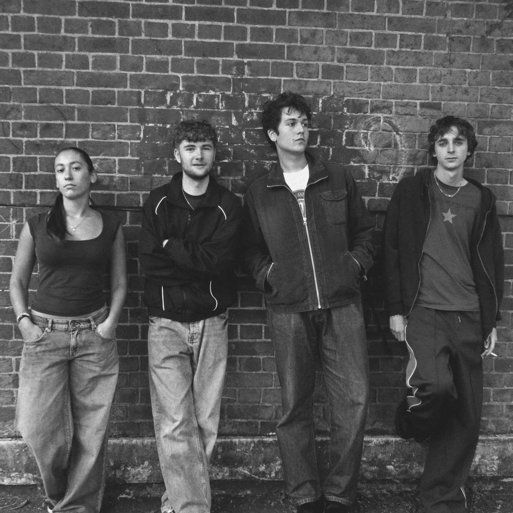 Four young people standing against a brick wall, with the two in the center leaning on the wall and the others standing with relaxed postures.