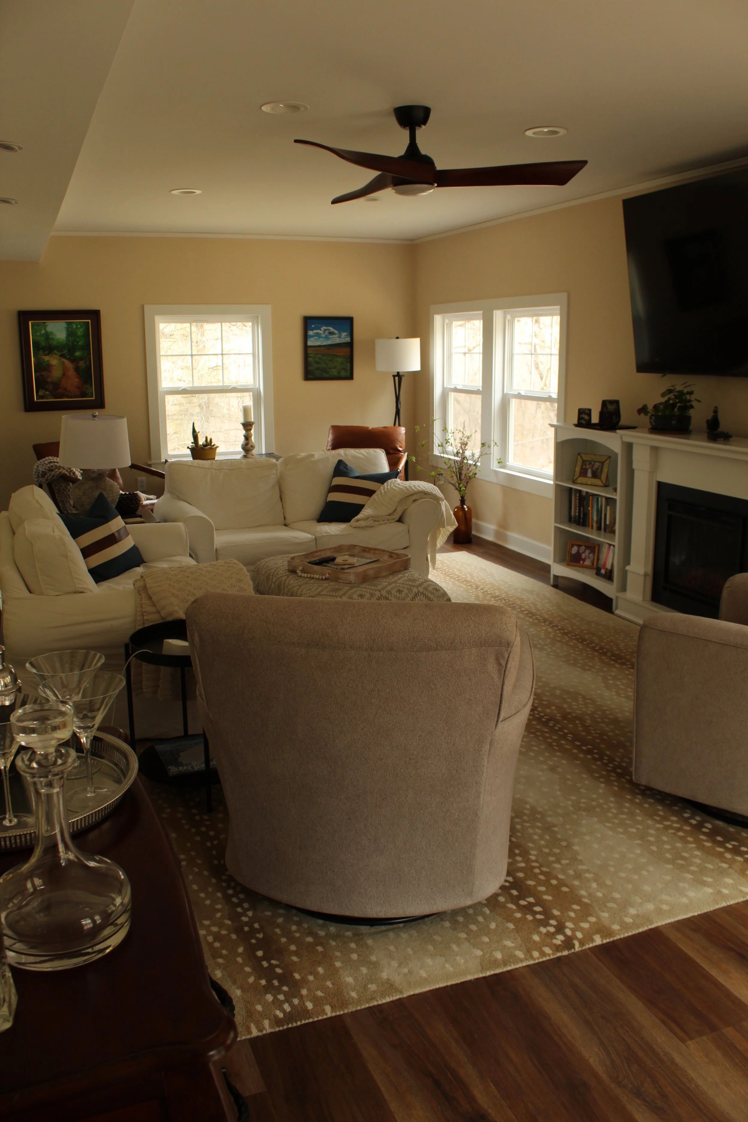Living room with cream-colored sofa, beige armchair, patterned rug, large windows, and wall-mounted TV.