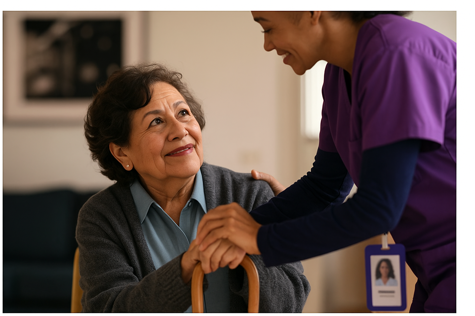 A friendly healthcare worker in purple scrubs holds hands and speaks with an elderly woman in a gray sweater, who is smiling and looking up at him.
