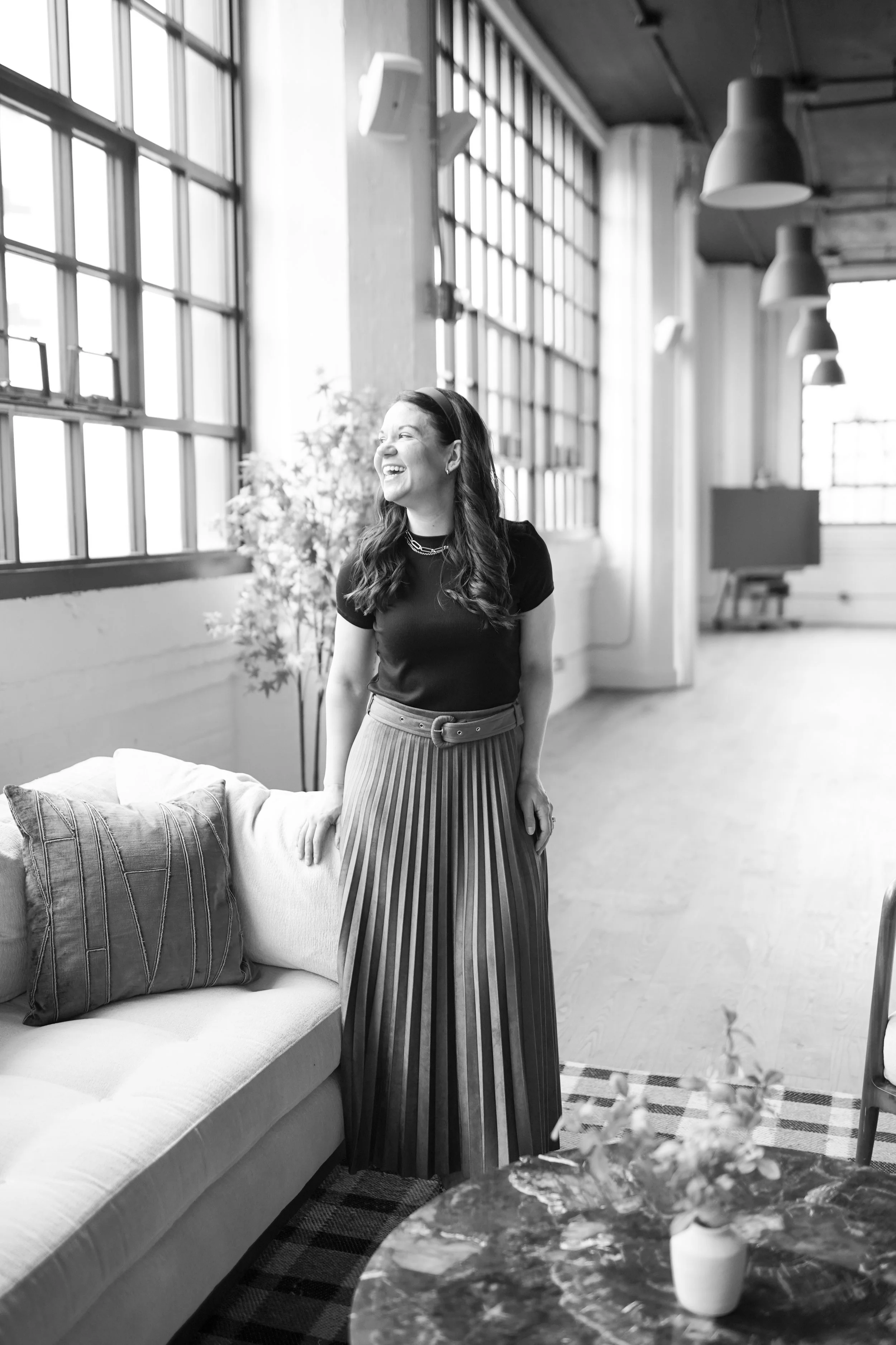 A woman with long, wavy hair and a big smile, wearing a dark top and a striped pleated skirt, standing next to a sofa in a bright, spacious room with large windows, plants, and hanging pendant lights.
