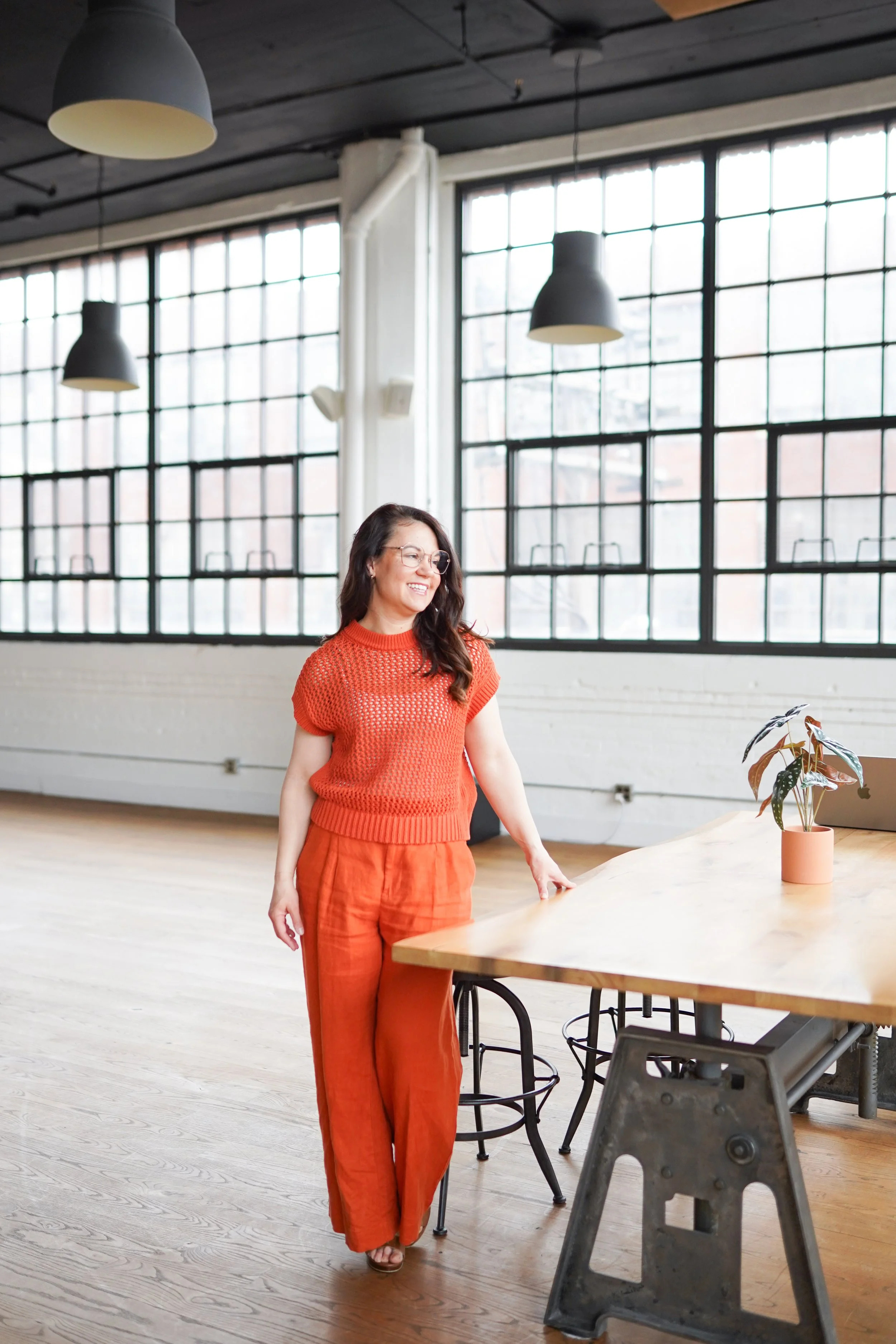 A woman in an orange outfit standing near a wooden table inside a bright, industrial-style room with large windows and hanging lights.