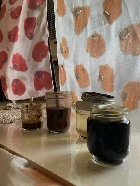 Four glass jars with dark liquids including a thickened madder root paste, iron water, a thickened osage-orange paste, and henna on a white surface in front of two of the poppy petal silk scarves in progress of being painted.