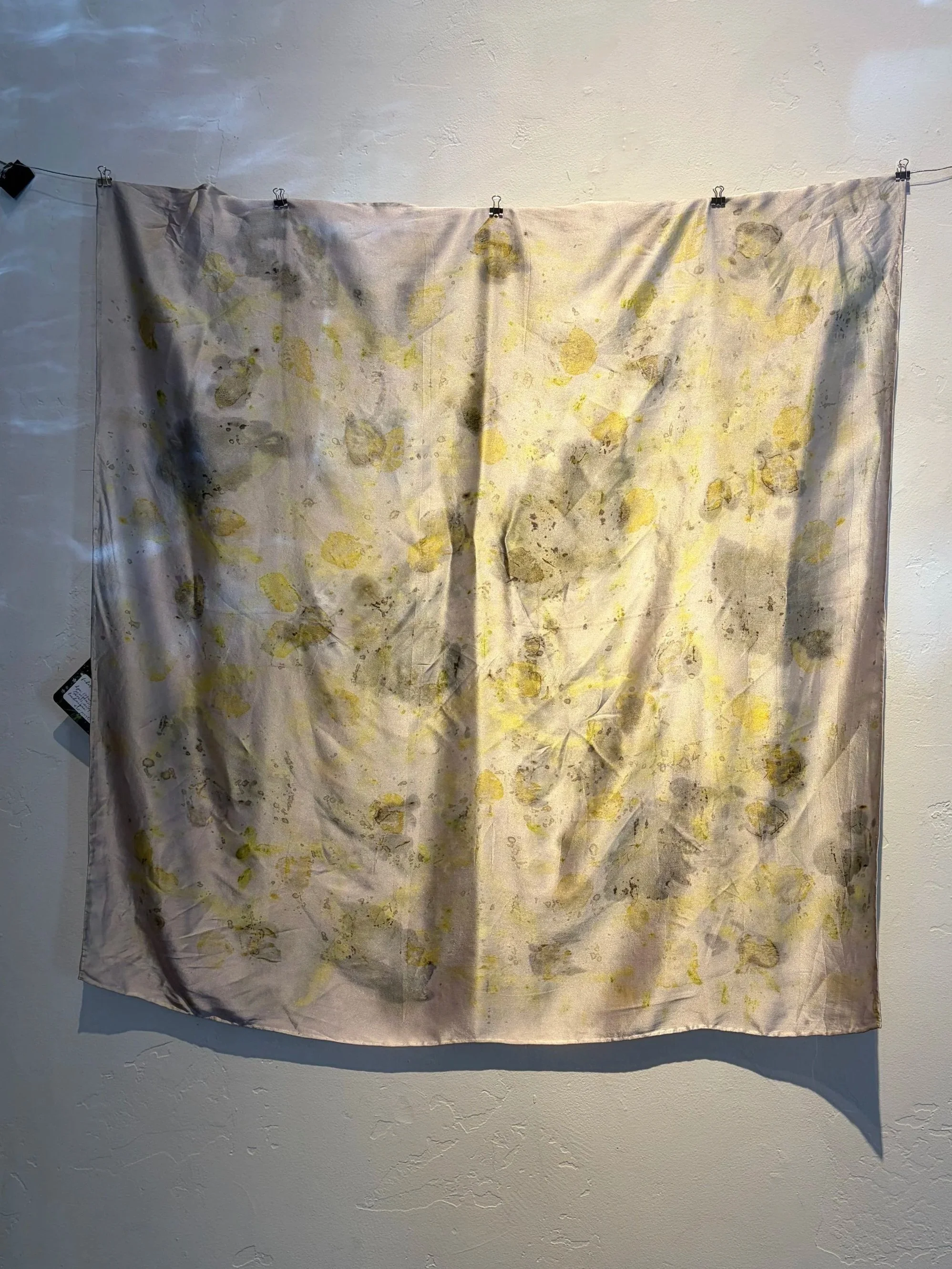 A piece of fabric hanging on a wire with clothespins, featuring abstract yellow and grayish-green patterns.