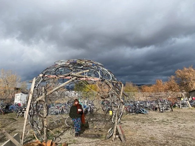 The artist behind the imprint collective bundled up with a cup of tea in a dystopian setting. She sits under a large outdoor sculpture made of old bicycle wheels, with a cloudy sky and other recycled bicycle parts strewn about.