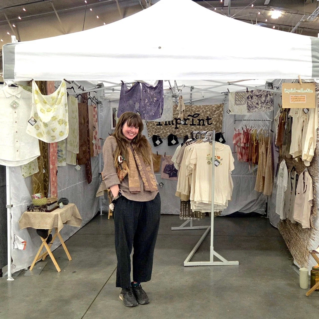 A woman standing inside a booth at a festival, smiling at the camera. The booth displays handmade and naturally dyed clothing, with the sign 'imprint' hanging on the back wall.