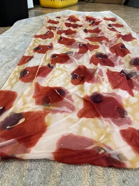 A sheer silk scarf with a pattern of red & black, depicting abstract poppy petal shapes, laying on a textured towel as the paint dries.