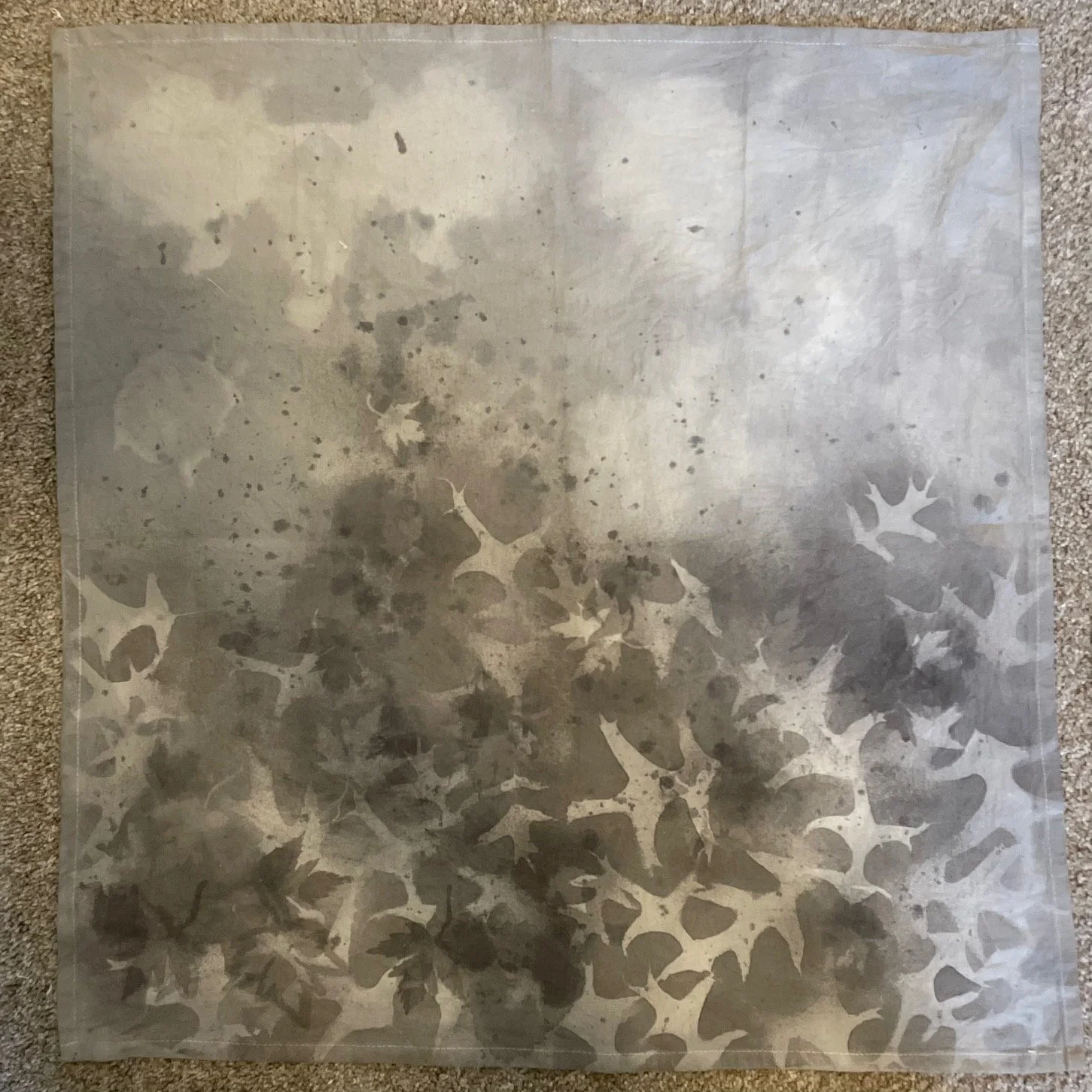 A soft textured kitchen towel with abstract leaf and splatter patterns overlayed, placed on a carpeted surface. Dyed using avocado pits and acorns from Iowa City, Iowa.