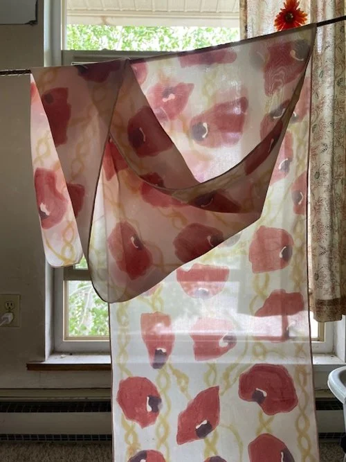 The hand-painted poppy petal habotai silk scarf hangs in front of a window, showcasing a pattern of red and black abstract shapes inspired by the poppy flowers. Dyed with madder root, iron, and minor batik detailing