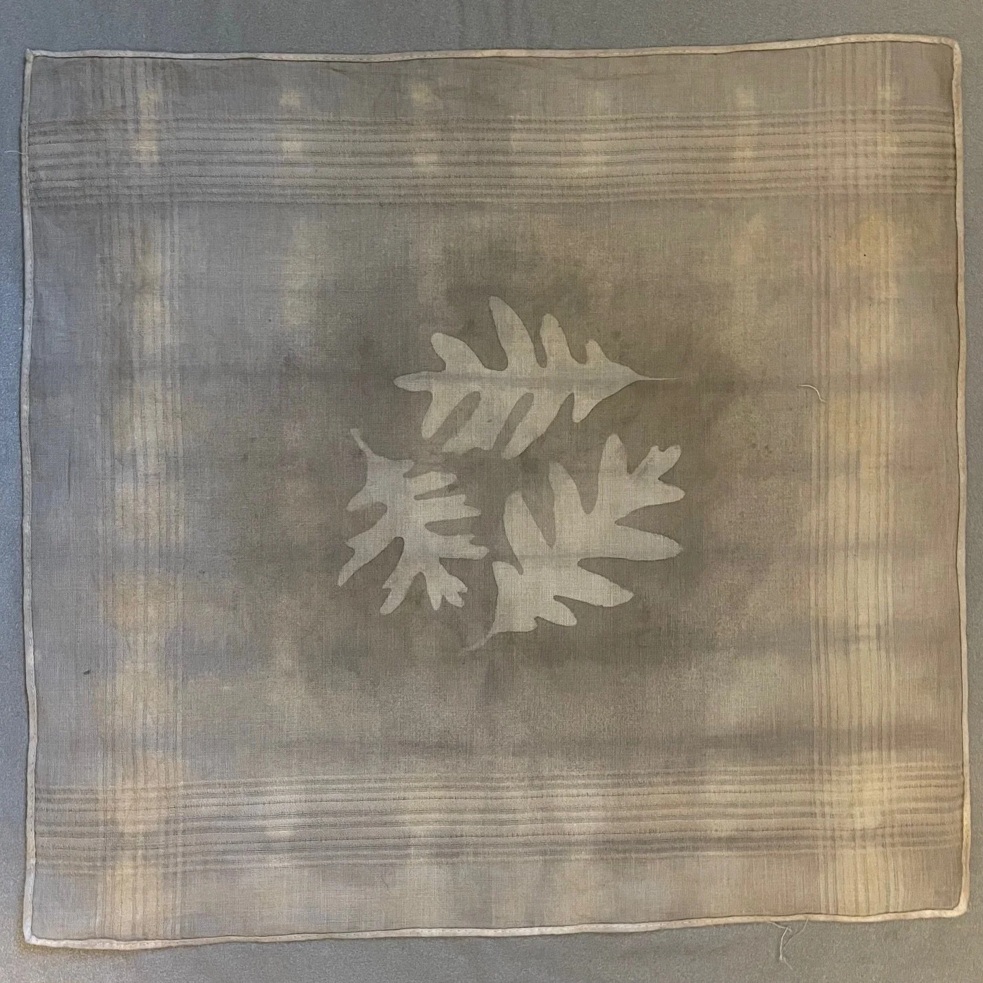 A handkerchief with a central cut-out design of three overlapping oak leaves created using a hand-made acorn dye, surrounded by a subtle striped and textured pattern created using traditional shibori techniques.