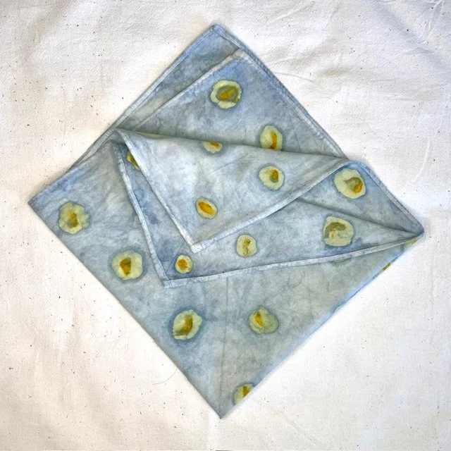 A naturally dyed 100% cotton bandana with a polk-a-dot pattern of eco-printed yellow marigolds and light circles created using the traditional batik method on a light blue indigo-dyed background, folded diagonally and placed on a white surface.