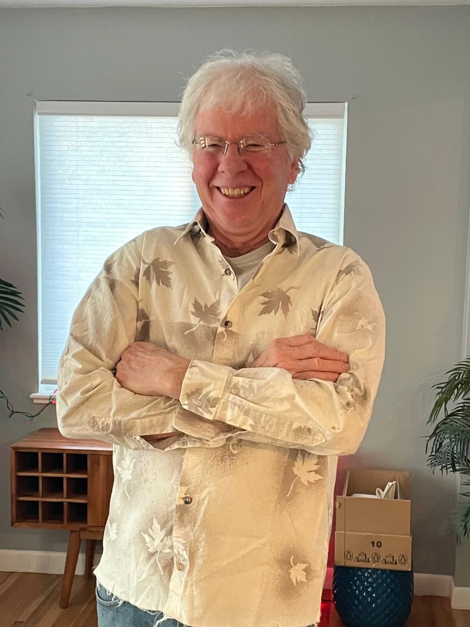 A smiling older man with white hair and glasses standing with arms crossed in a room with a window, a wooden cabinet, a plant, and a box of items. He wears a hand-made and hand-dyed, naturally-dyed prototype that used a hand-made acorn dye, stencils