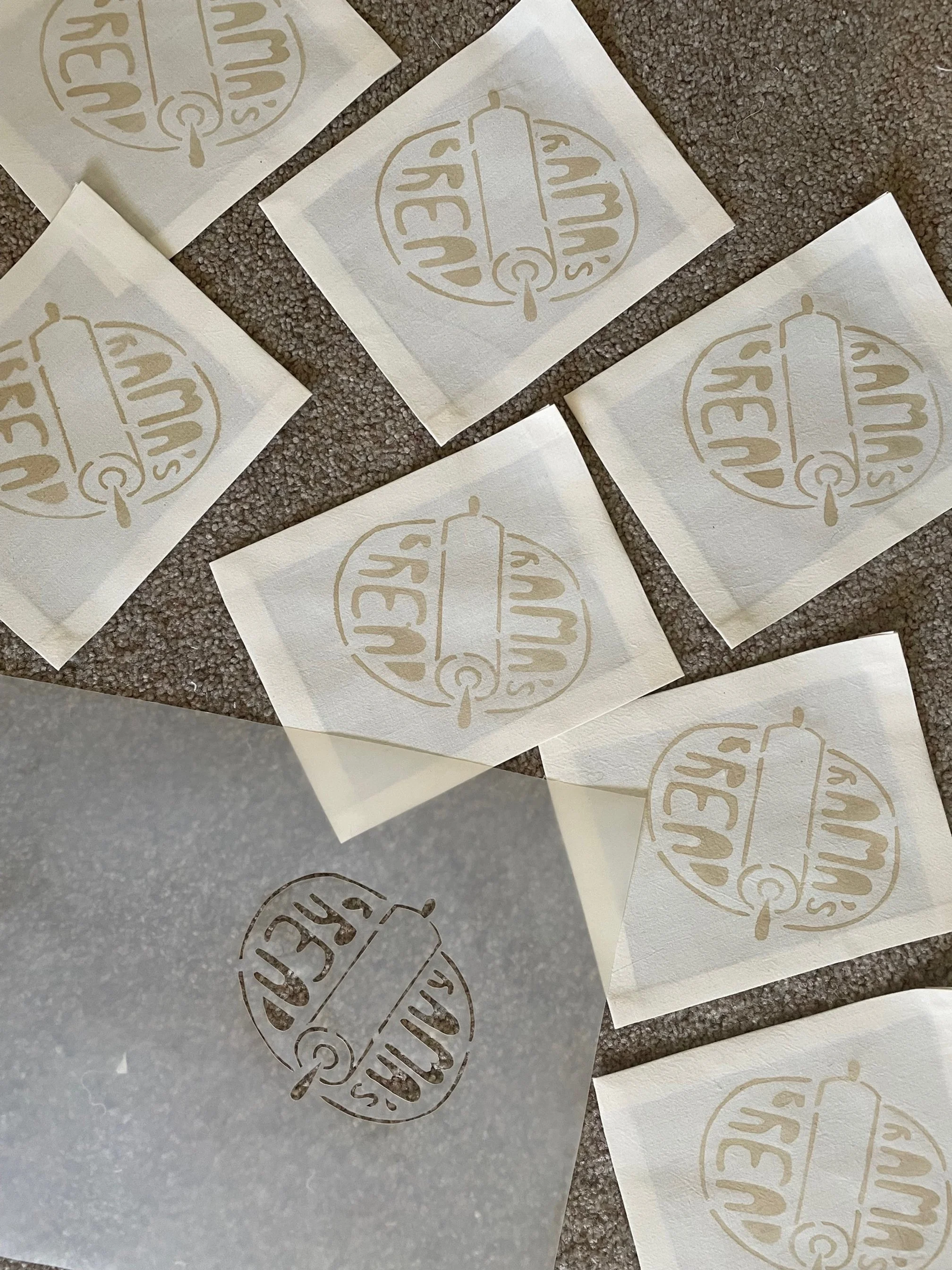 Multiple sheets of fabric with a circular logo featuring the text "Rama's Bread" and a dough with a rolling pin illustration created from a stencil and hand-made acorn dye. Next to these are the stencil used to create the designs.