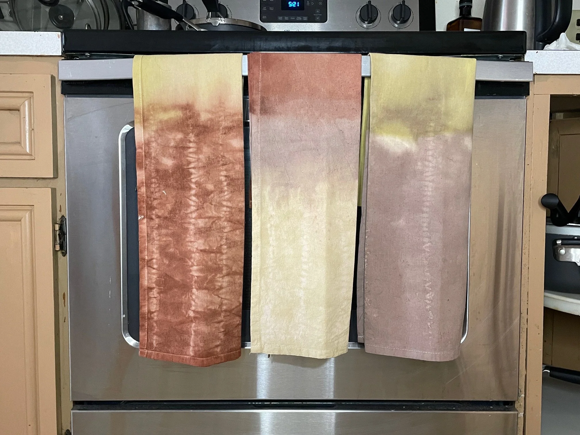 Three naturally-dyed kitchen towels in shades of brown, yellow, and beige hanging over a stove in a kitchen. These colors are naturally created using cutch and henna