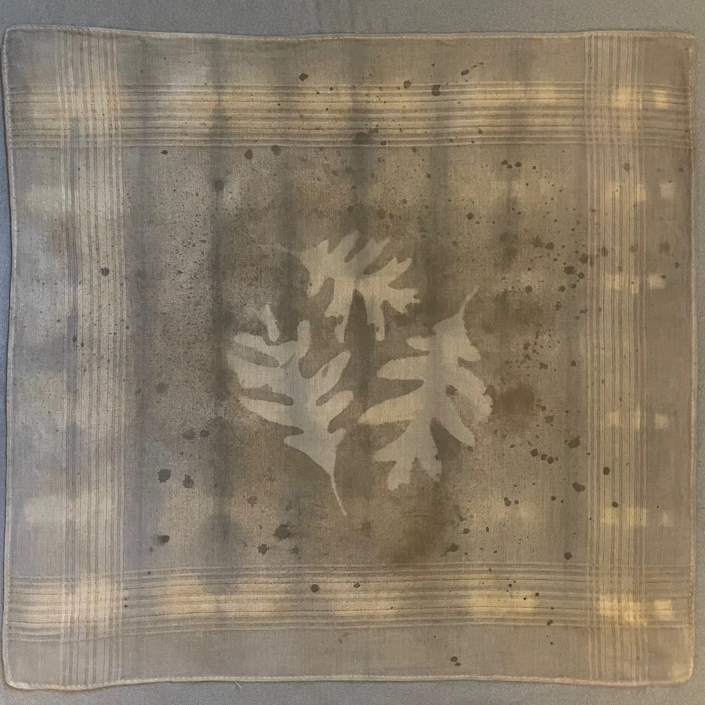 A handkerchief with a central cut-out design of three overlapping oak leaves created using a hand-made acorn dye, surrounded by a subtle striped and textured pattern created using traditional shibori techniques.