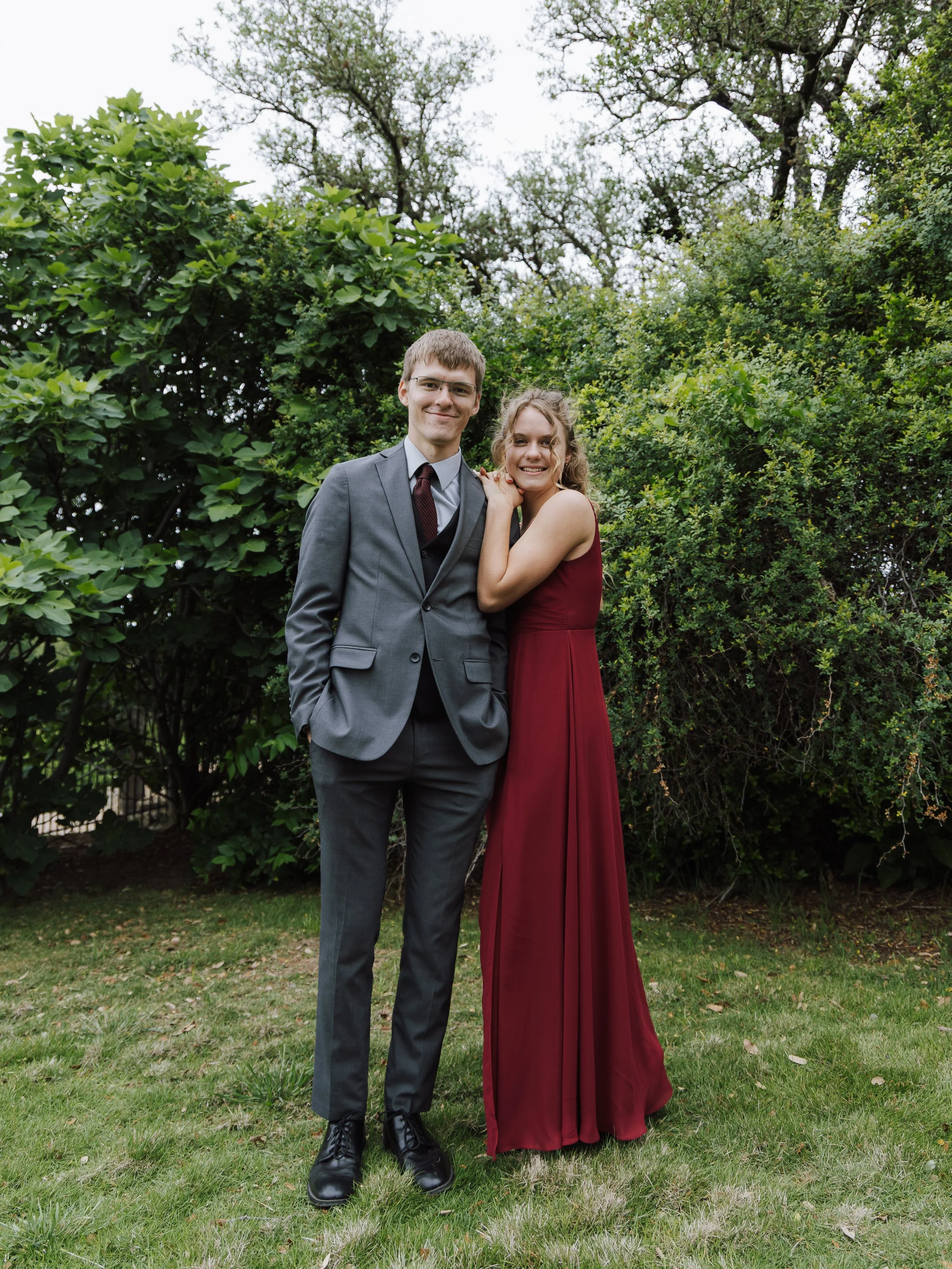A young couple standing on grass in front of bushes and trees. The man is wearing a gray suit and glasses, while the woman is in a long red dress. They are smiling and posing together outdoors.