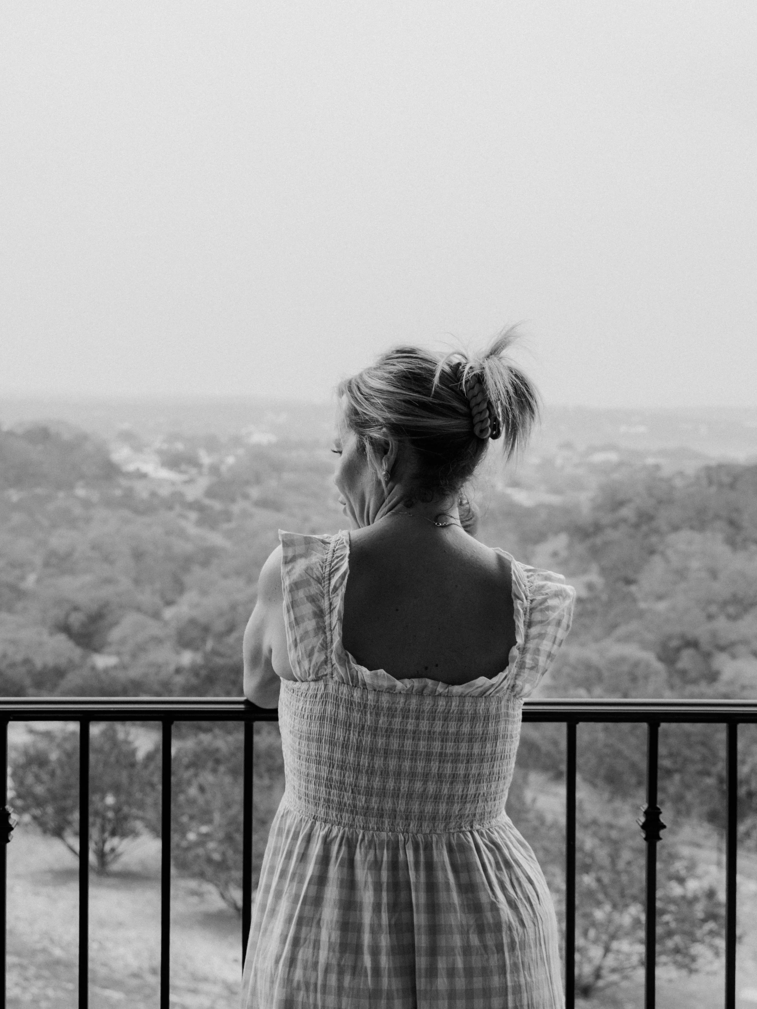 A woman with her back to the camera, standing by a fence, overlooking a landscape with trees and hills.