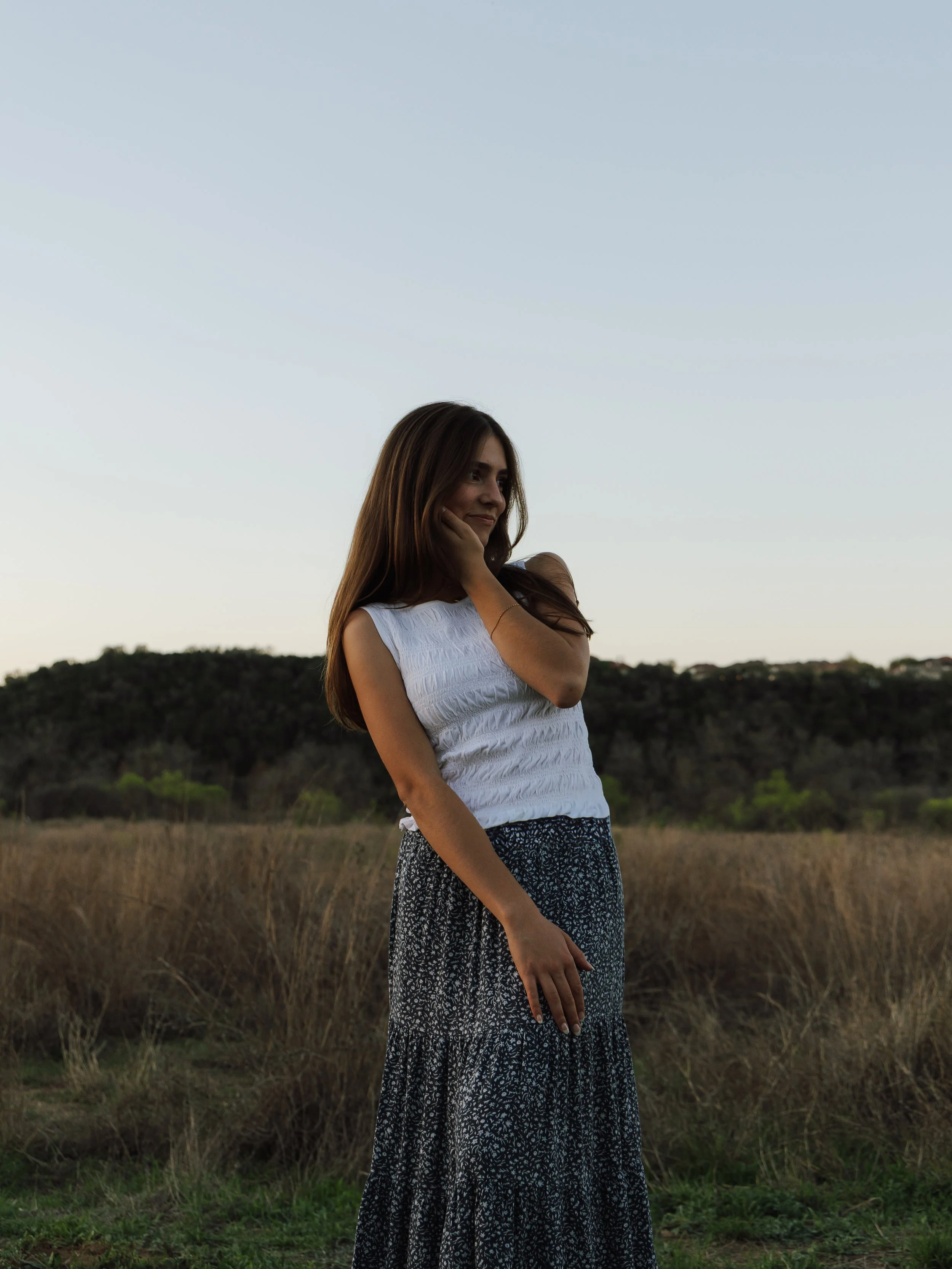 A woman with long brown hair standing in a field with dry grass and trees in the background during dusk. She is wearing a white sleeveless top and a long patterned skirt, looking thoughtfully to the side with her hand on her chin.