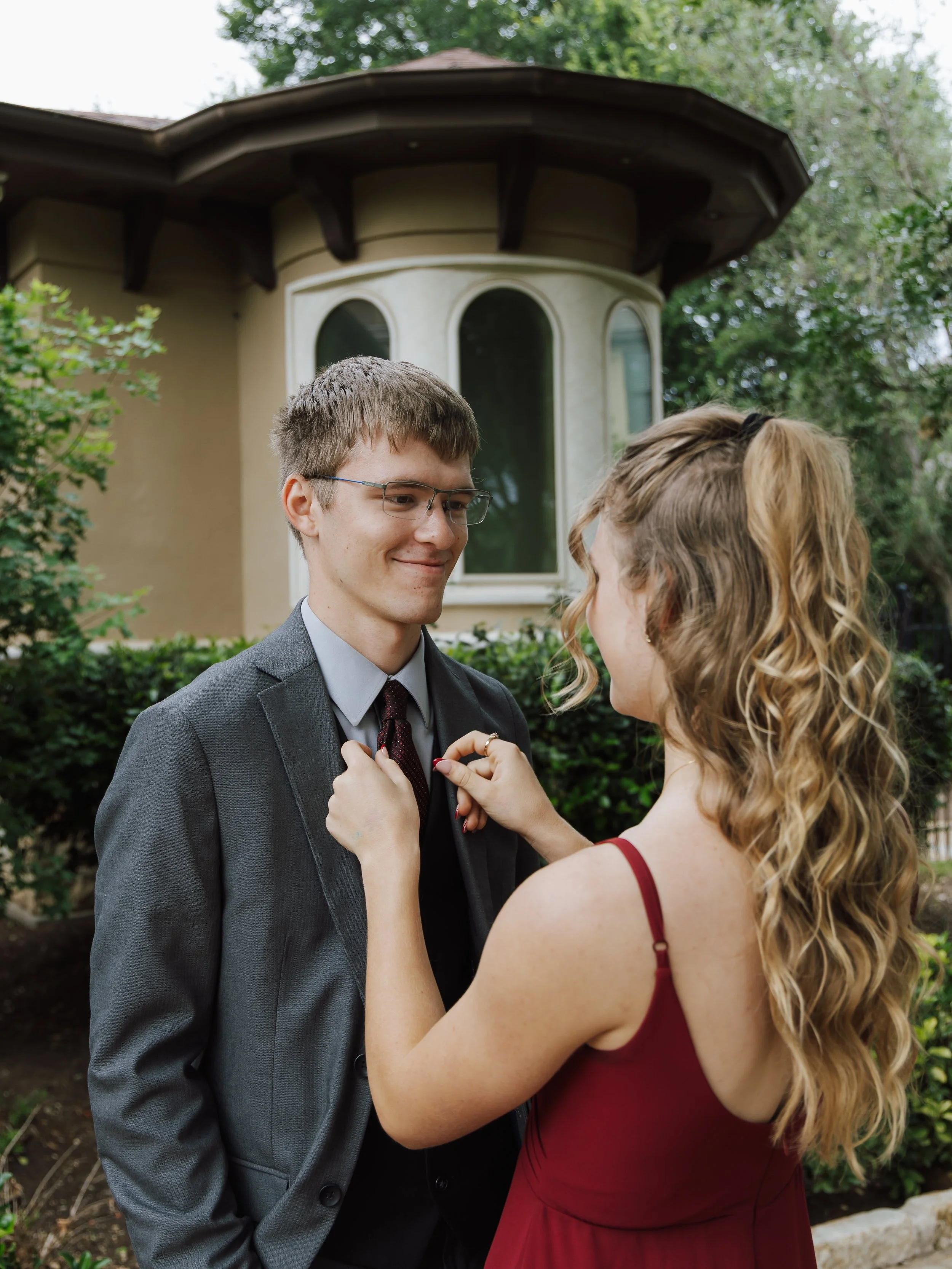 A woman pinning a boutonniere on a smiling young man dressed in a suit outdoors in front of a house with bay windows, trees, and bushes.