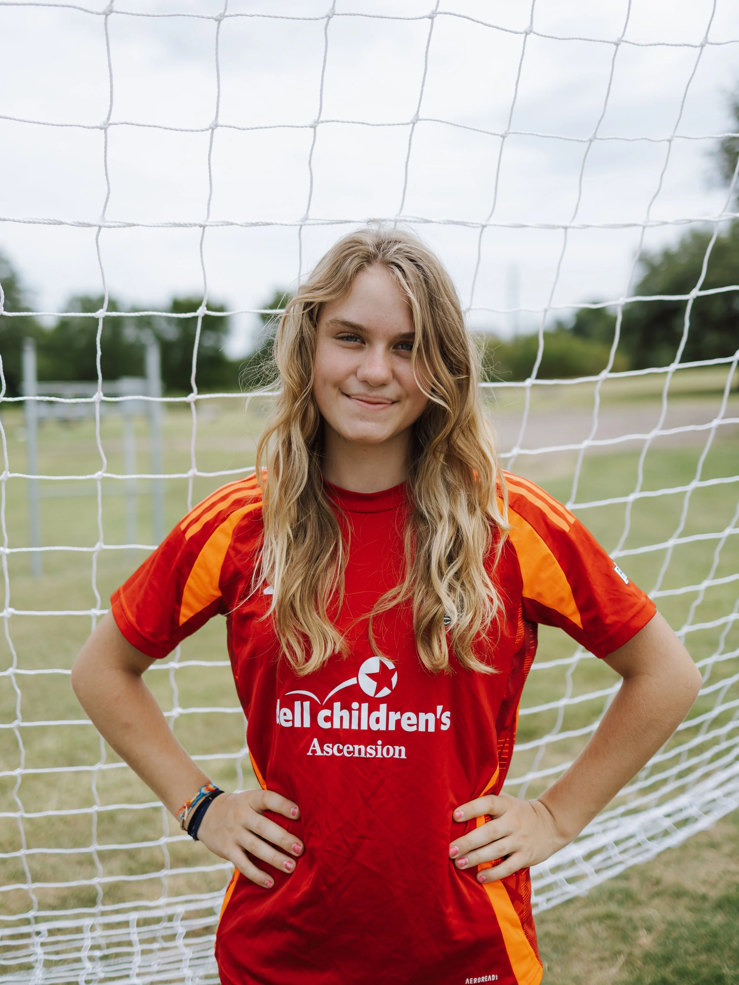 A young woman with long blonde hair standing smiling in front of a soccer goal net on a field, wearing a red and orange soccer jersey with the 'tell Children's Ascension' logo.