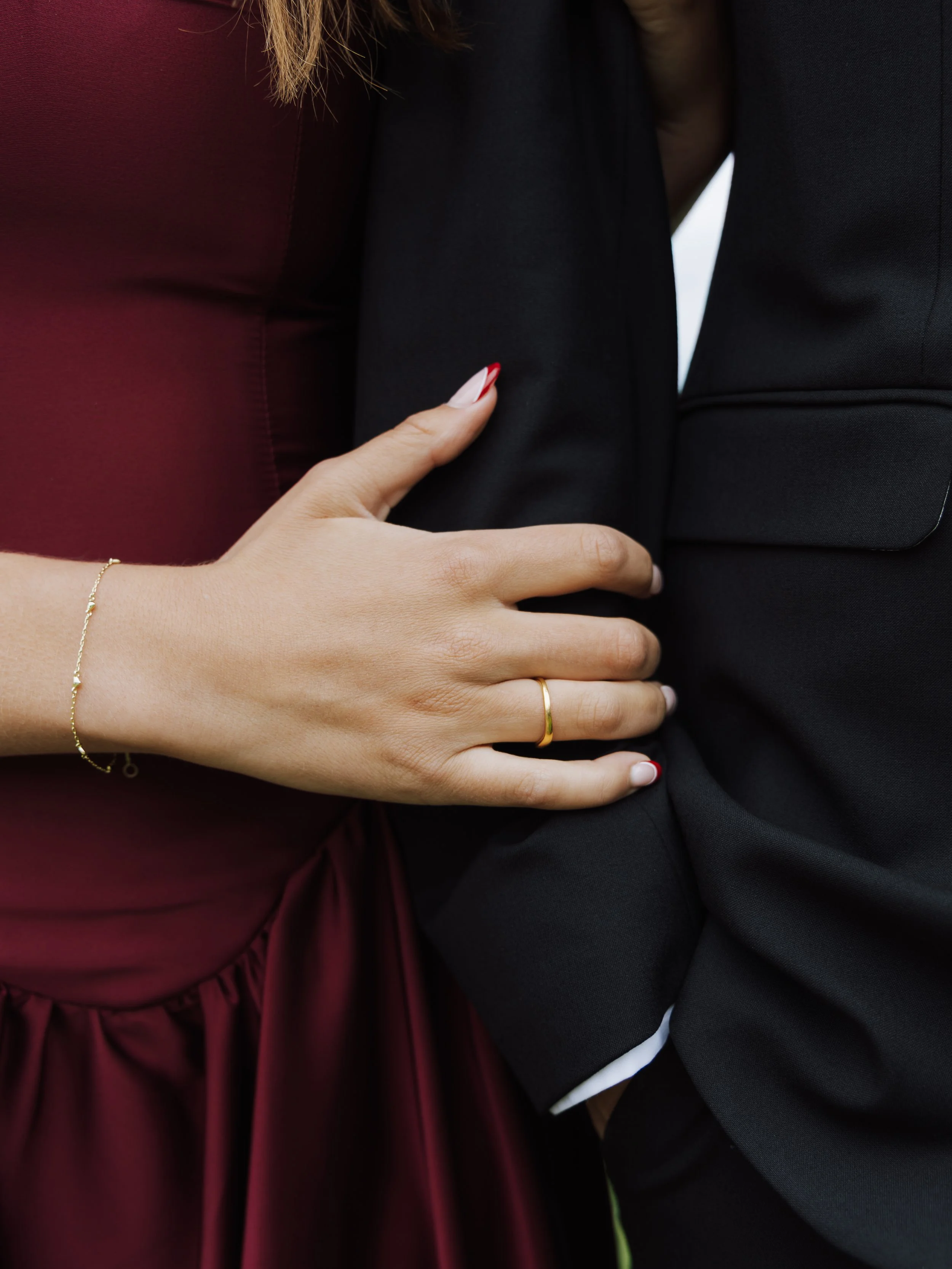 Close-up of a woman's hand resting on a man's chest, with both dressed in formal attire, featuring a gold ring and bracelet.