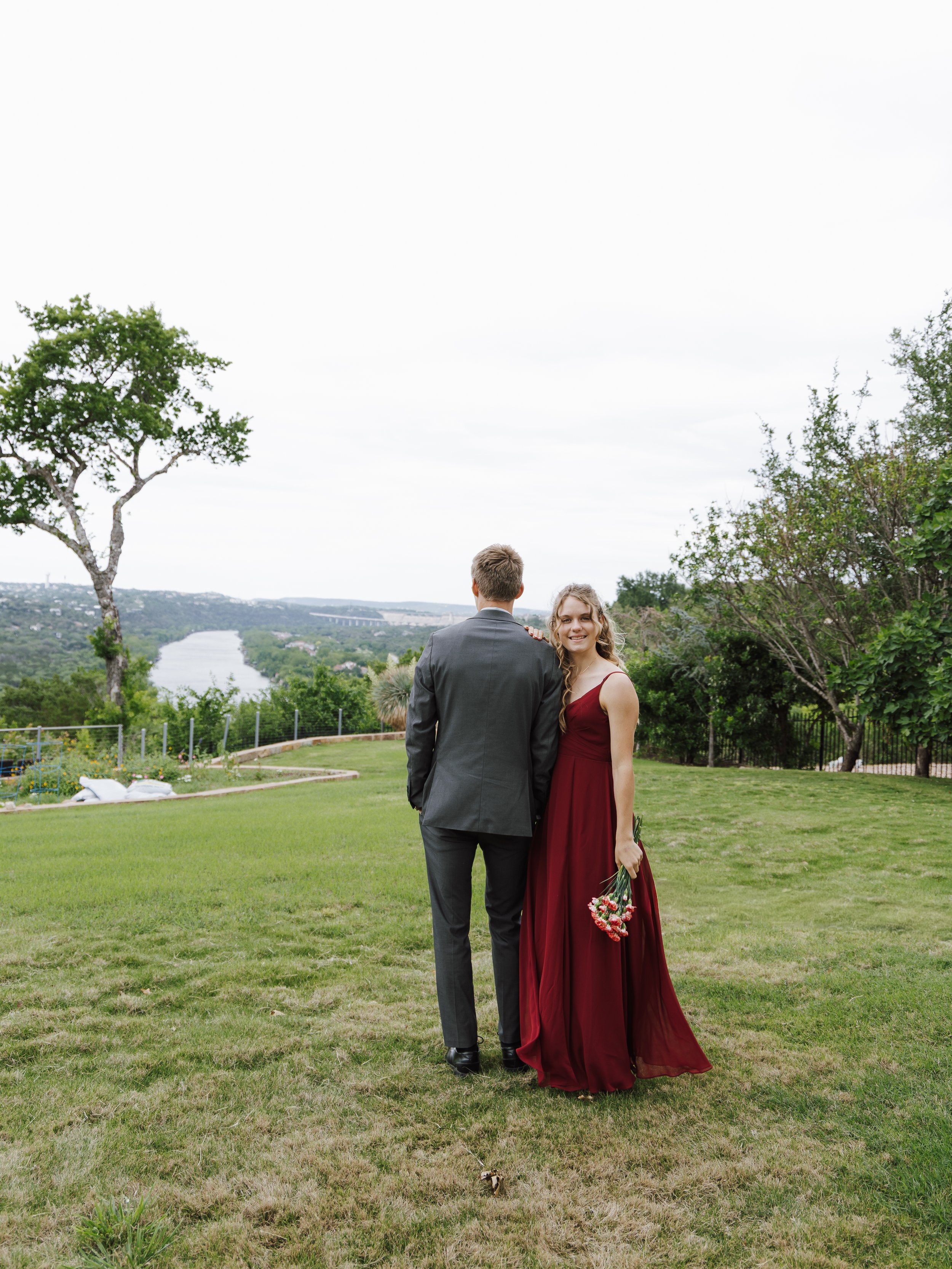 Couple standing outside on a grassy hill, with a river, bridge, and trees in the background, during daytime. The woman is holding a bouquet of pink flowers and smiling at the camera, wearing a red dress. The man is wearing a gray suit and has his bac