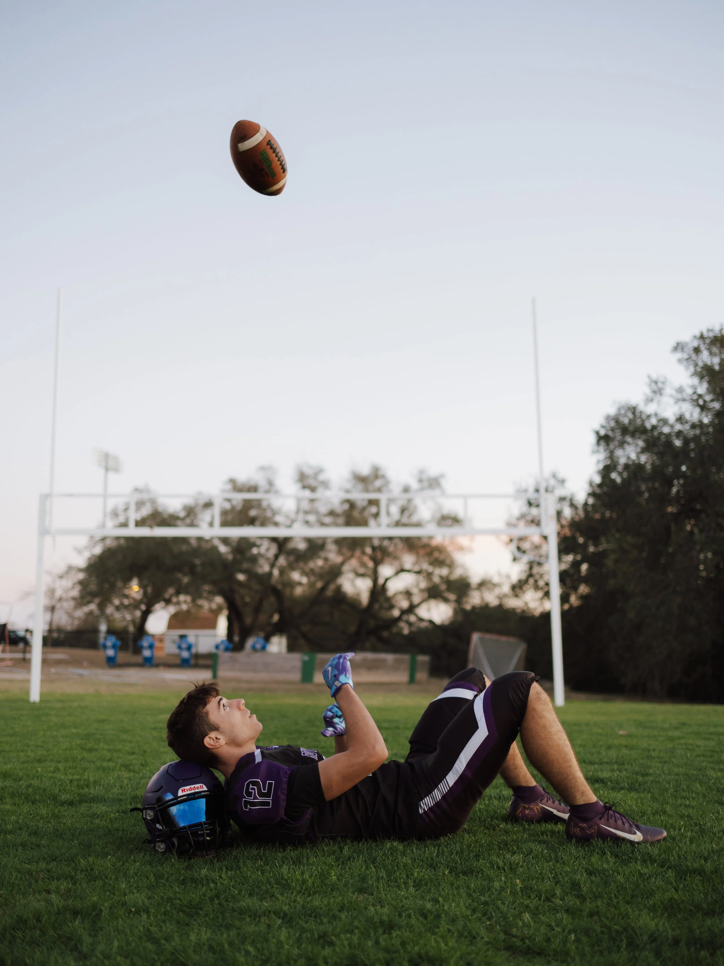 A football player lying on the grass field after a game or practice, with his helmet beside him, and a football flying through the air above him, while other players are visible in the background.