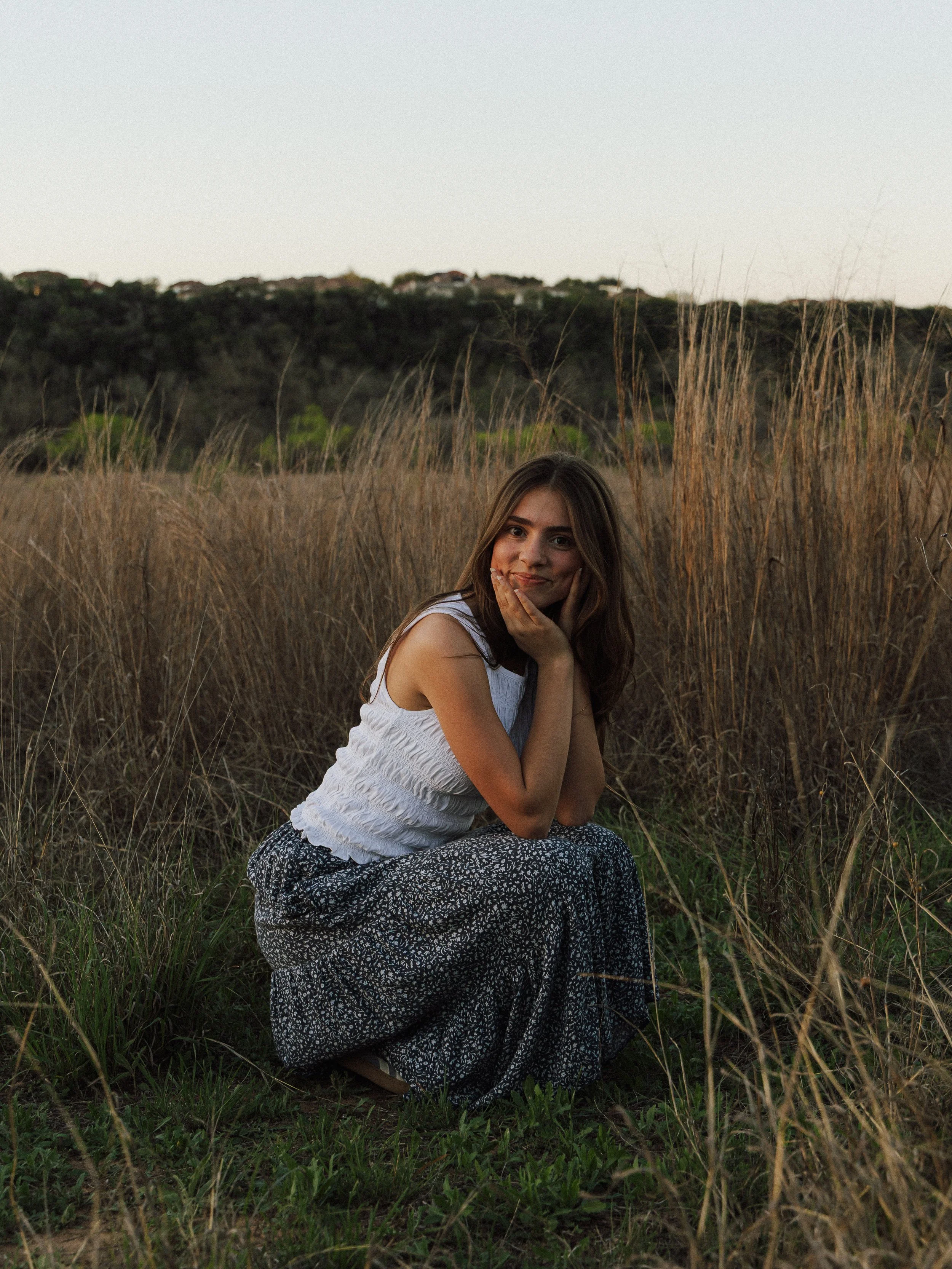 A young woman in a white sleeveless top and patterned skirt kneels in tall grass, smiling with her hands on her cheeks in an outdoor field during sunset.