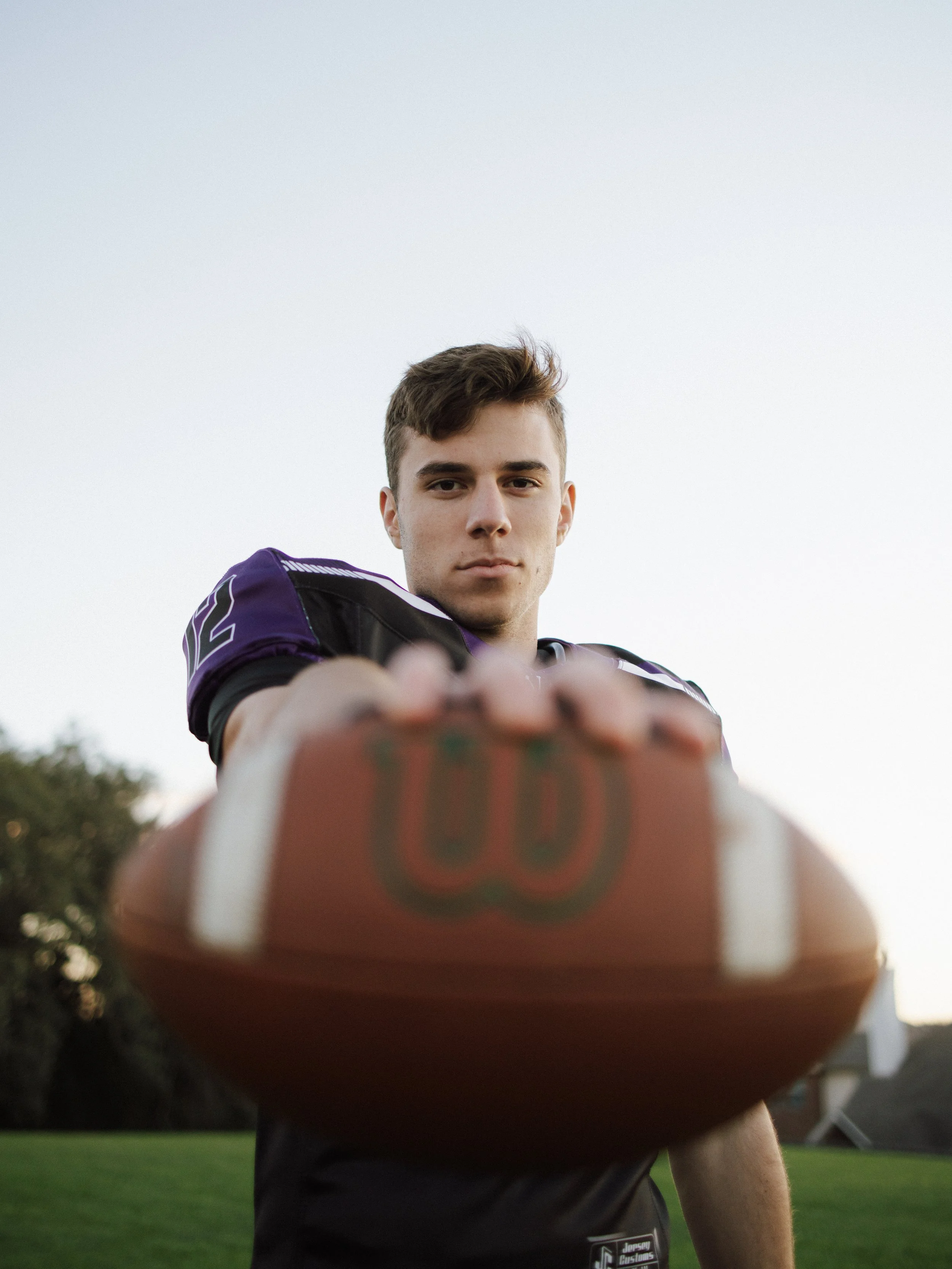 A young male football player in a purple jersey, looking directly at the camera, is holding a football close to the camera, with the focus on the football.