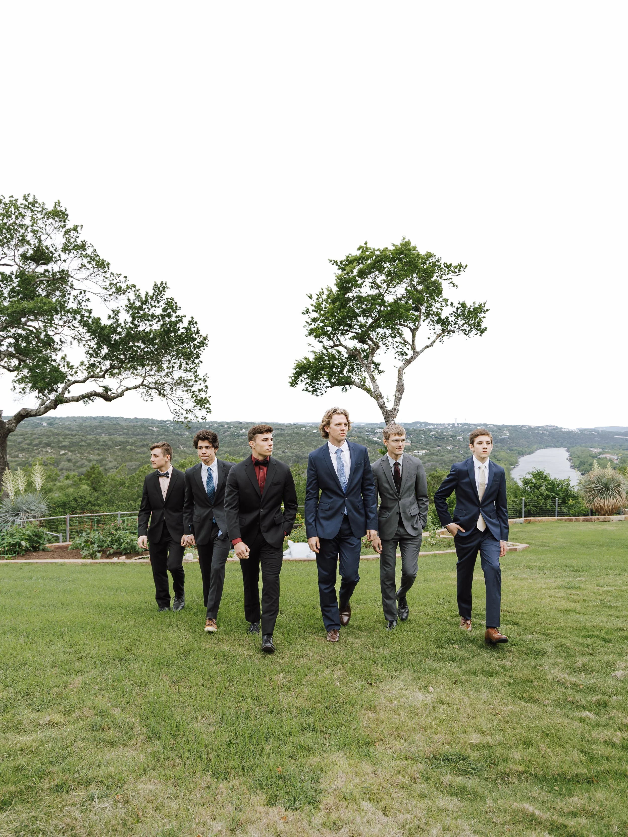 Six young men in suits walking on a grassy lawn with trees, a river, and a cityscape in the background.