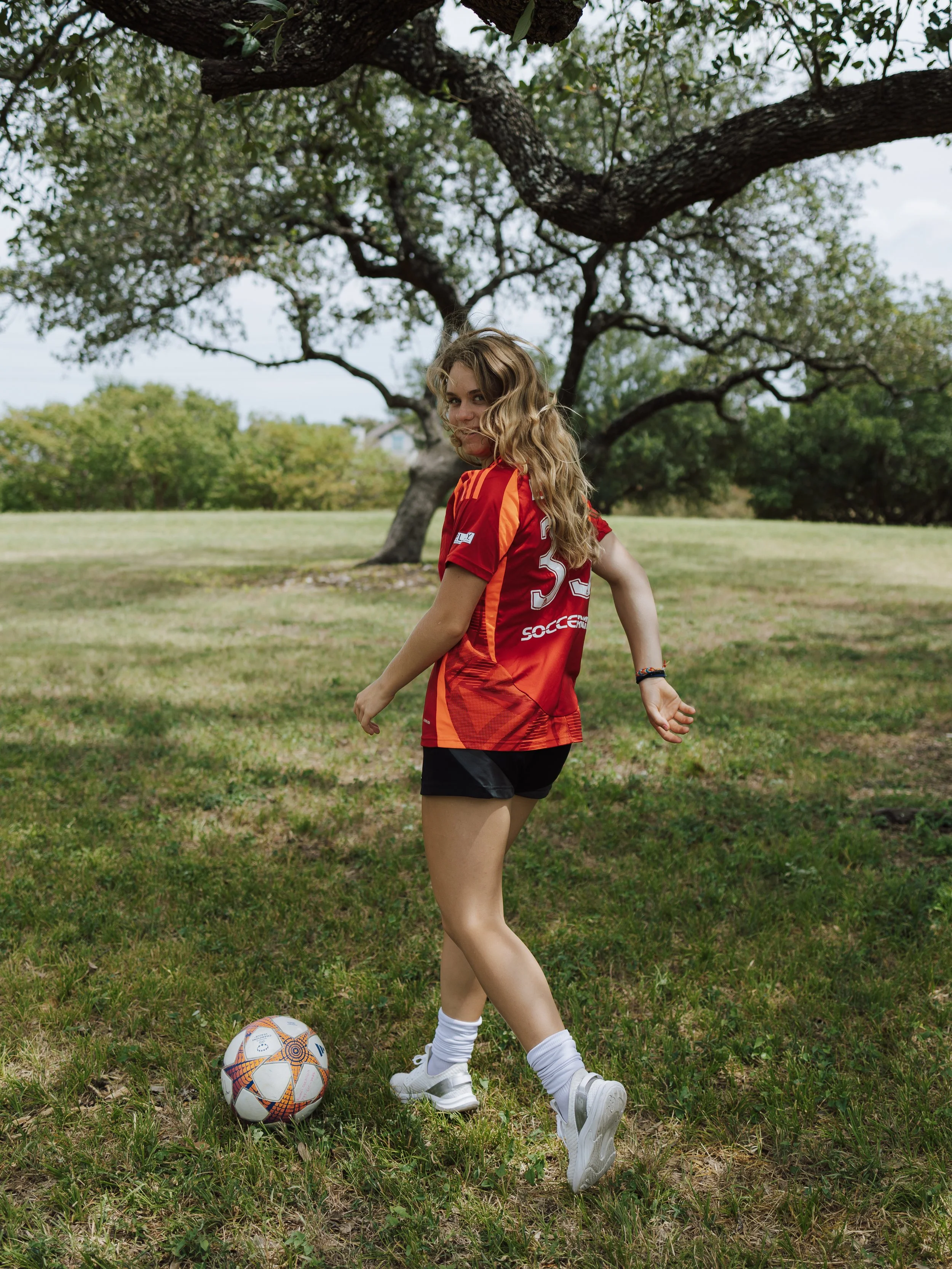 A young woman with blond curly hair, wearing a red soccer jersey, black shorts, white socks, and white sneakers, standing on grass with a soccer ball at her feet under a large tree in a park.