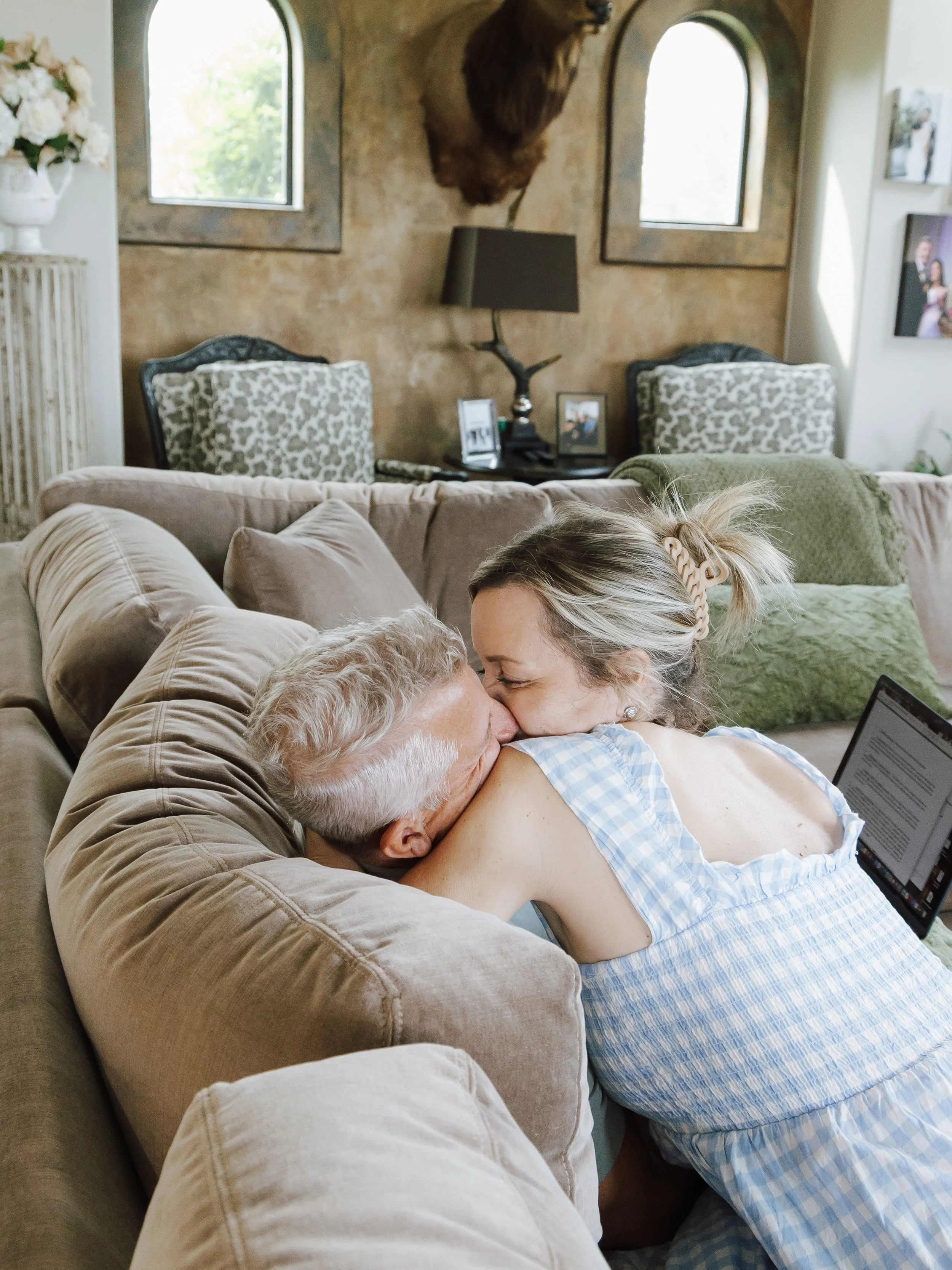 A woman and an older man sharing a kiss on a beige couch in a cozy living room with patterned cushions, a wall-mounted bear head, and pictures on the wall.