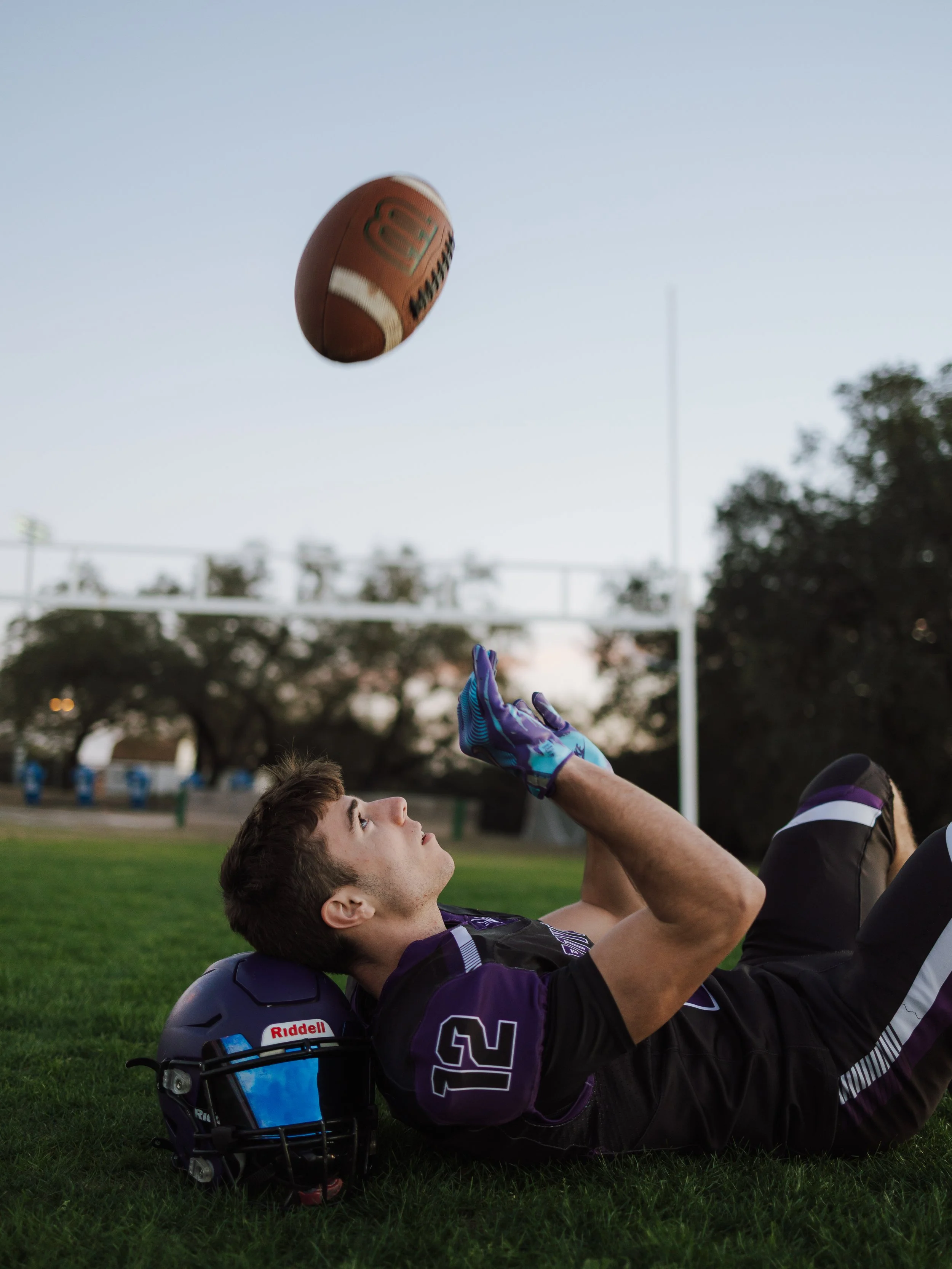A football player lying on the field, reaching up to catch a football. The player is wearing a football helmet and football uniform, with trees and goal posts visible in the background.