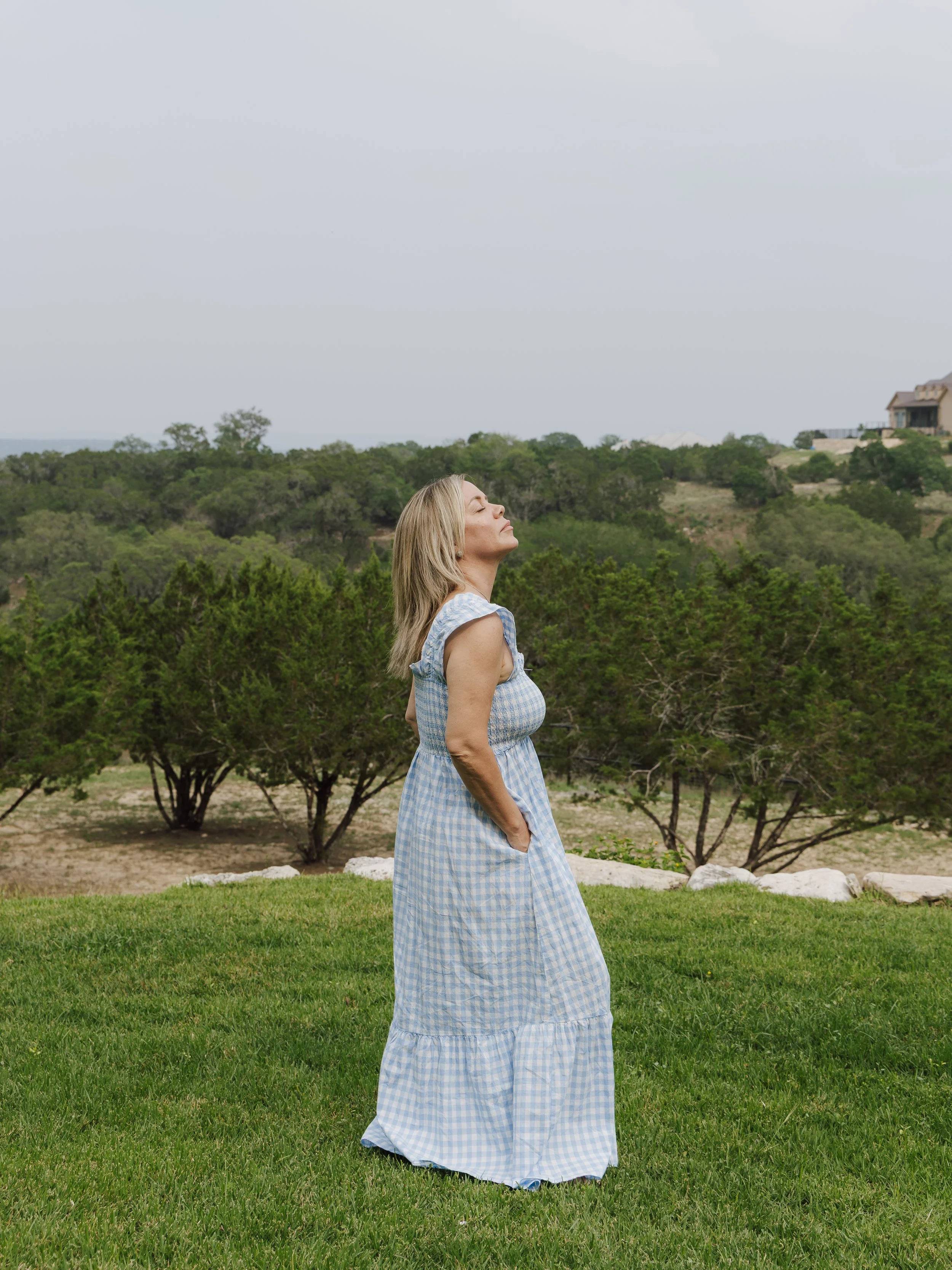 A woman with blonde hair wearing a blue and white checkered dress, standing outdoors with her eyes closed and face tilted slightly upward, surrounded by green grass and trees, with houses and hills in the background under a cloudy sky.