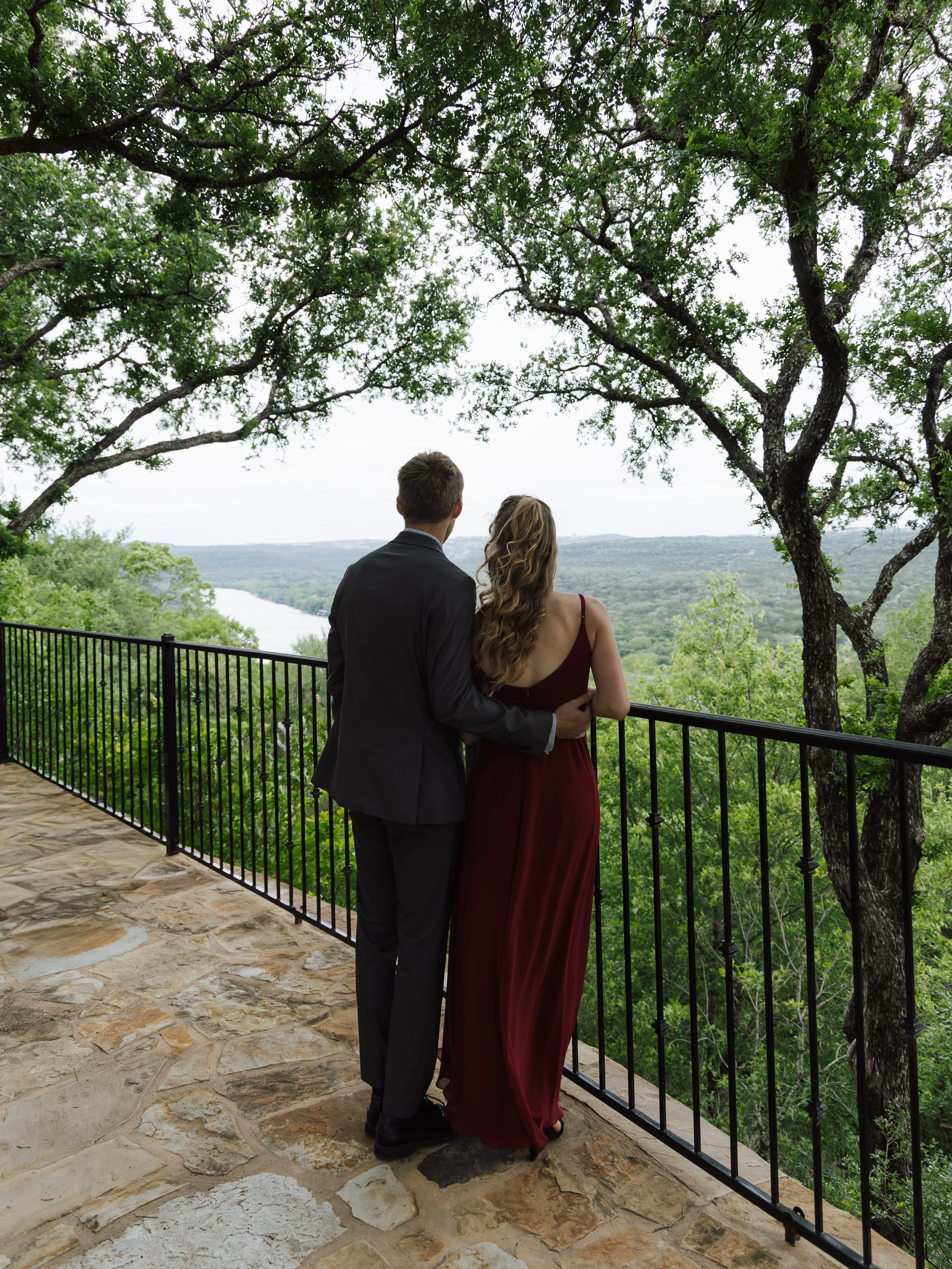 A couple dressed in formal attire standing close together on a balcony overlooking a scenic landscape with trees and a river, surrounded by lush greenery and large trees.