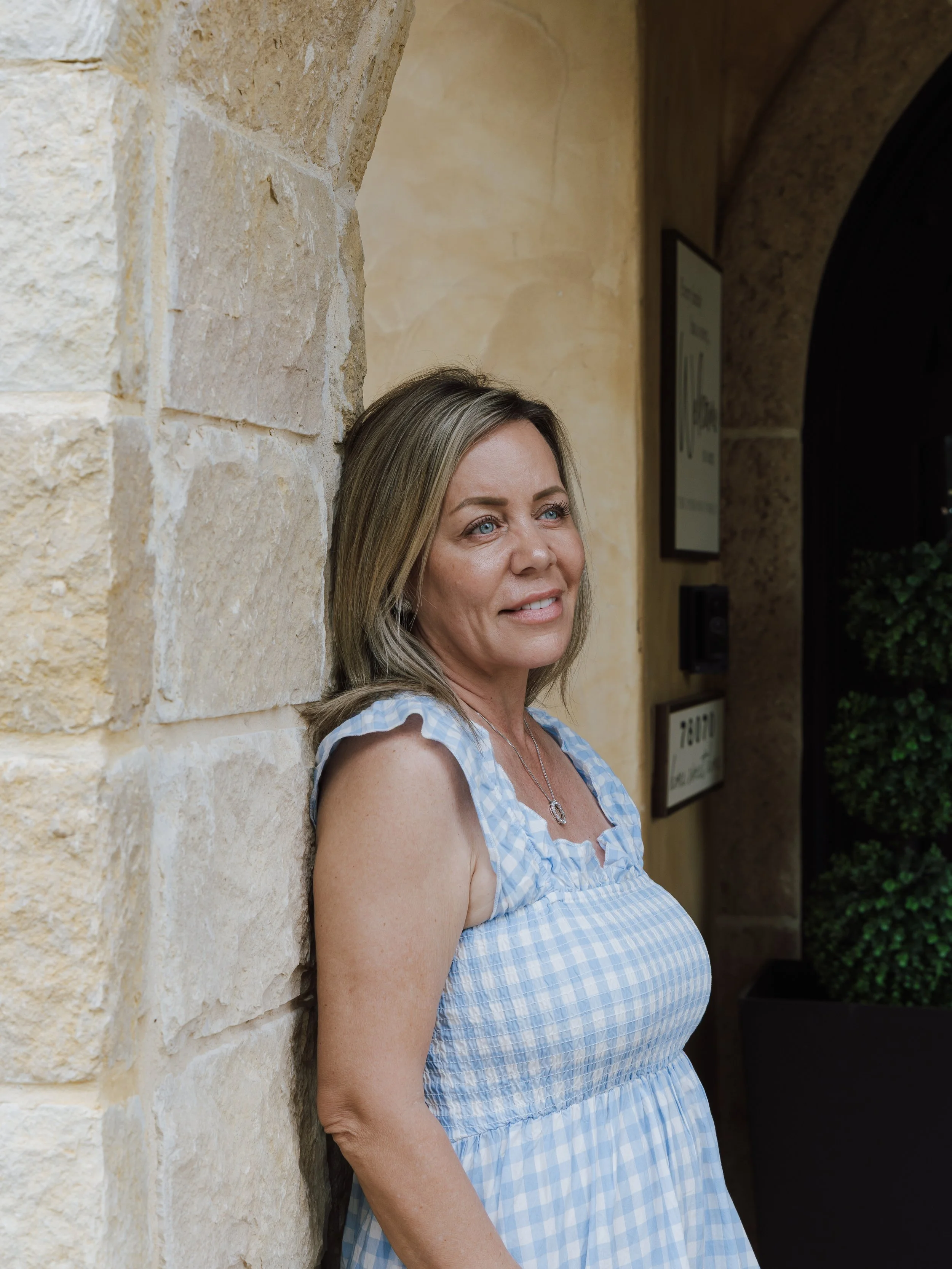A woman with blonde hair and blue eyes leaning against a beige stone wall, wearing a blue and white checkered sleeveless dress.