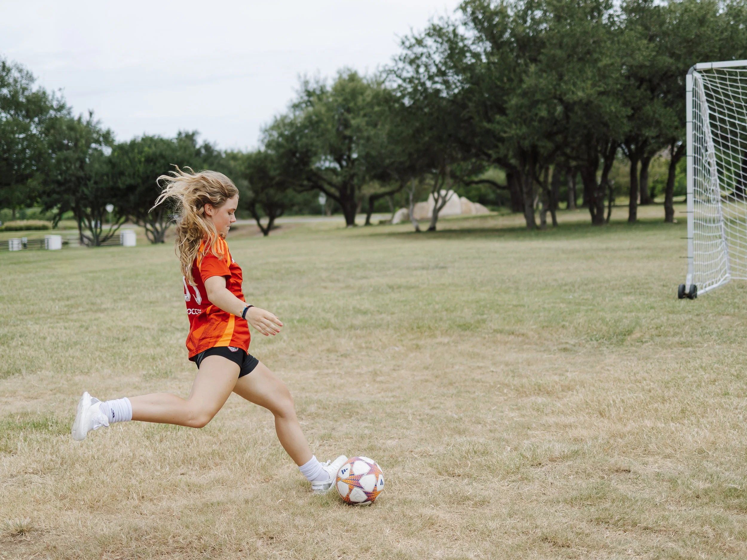 A young girl in athletic clothing kicks a soccer ball on a grassy field with trees and an outdoor soccer goal in the background.