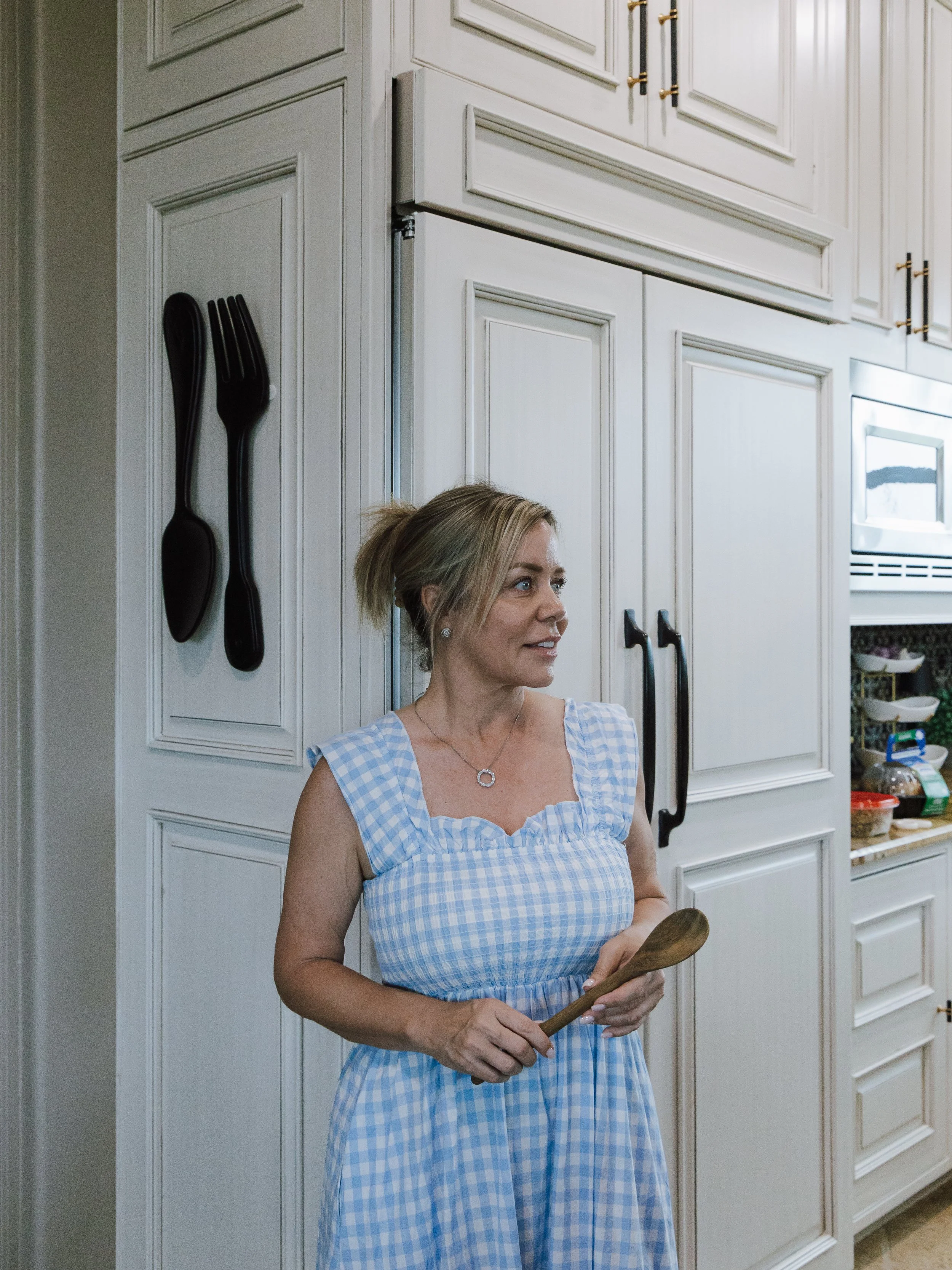 A woman with blonde hair tied back, wearing a blue and white checkered dress, holding a wooden spoon, standing in a kitchen with white cabinets, a microwave, and various food items on the counter.